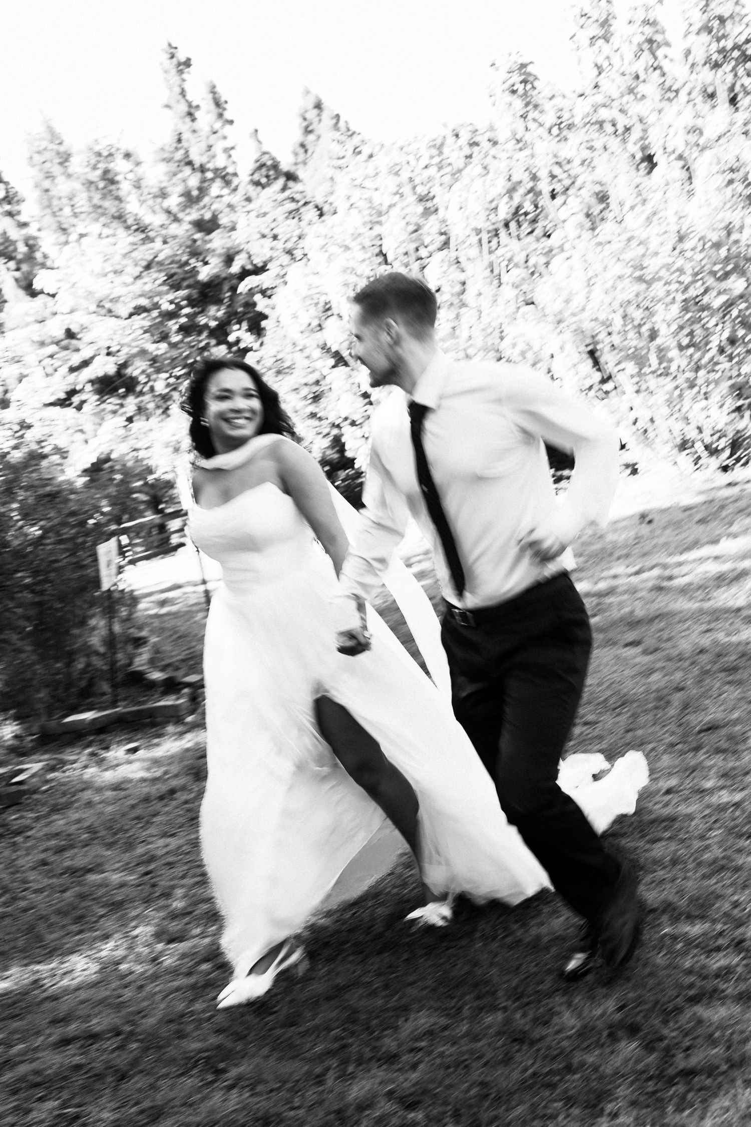 Black and white photo of a joyful bride and groom running together outdoors on their wedding day.
