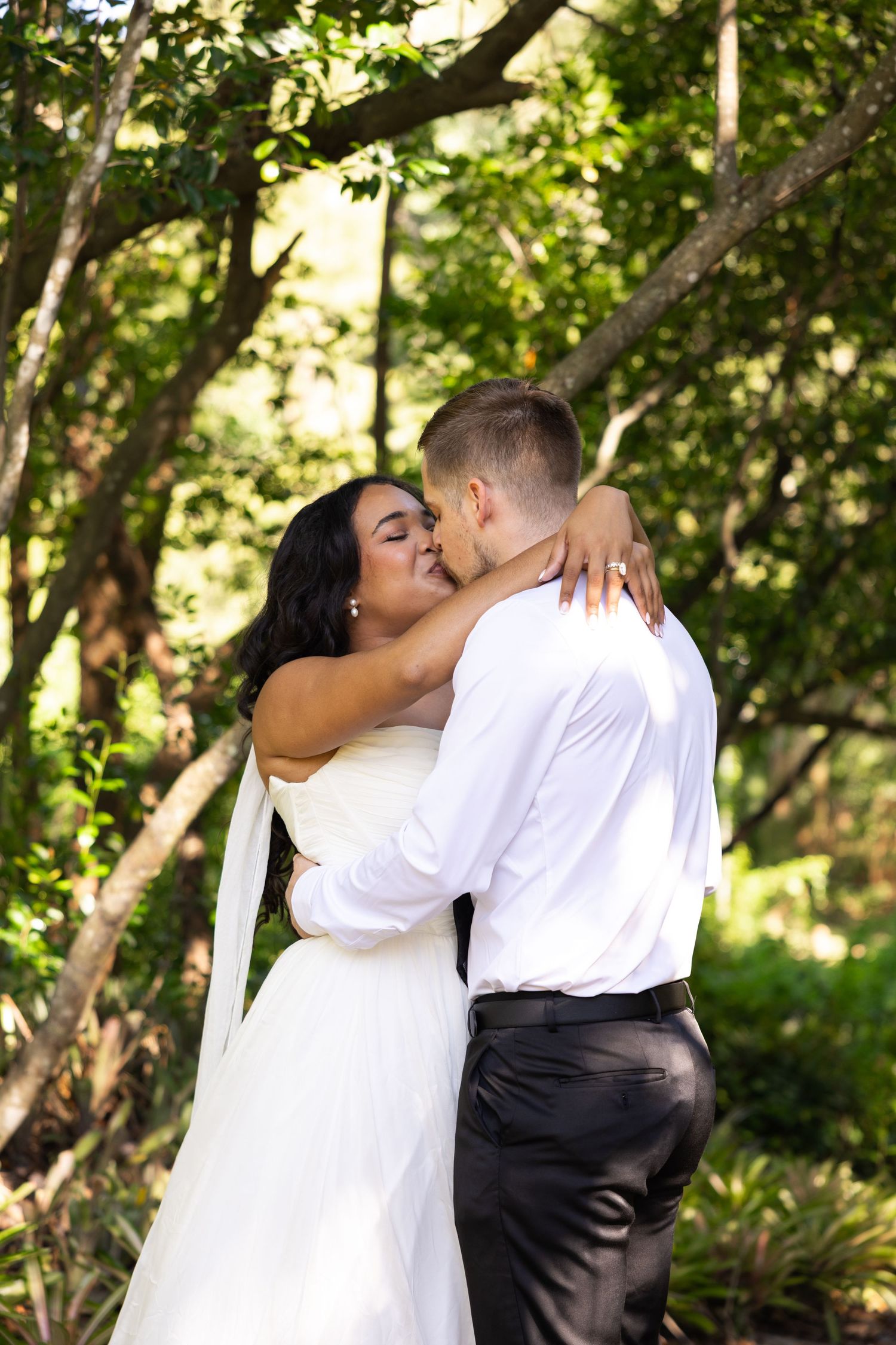 Newlywed couple sharing a romantic kiss outdoors, bride in white gown with veil, groom in white shirt, lush green trees.