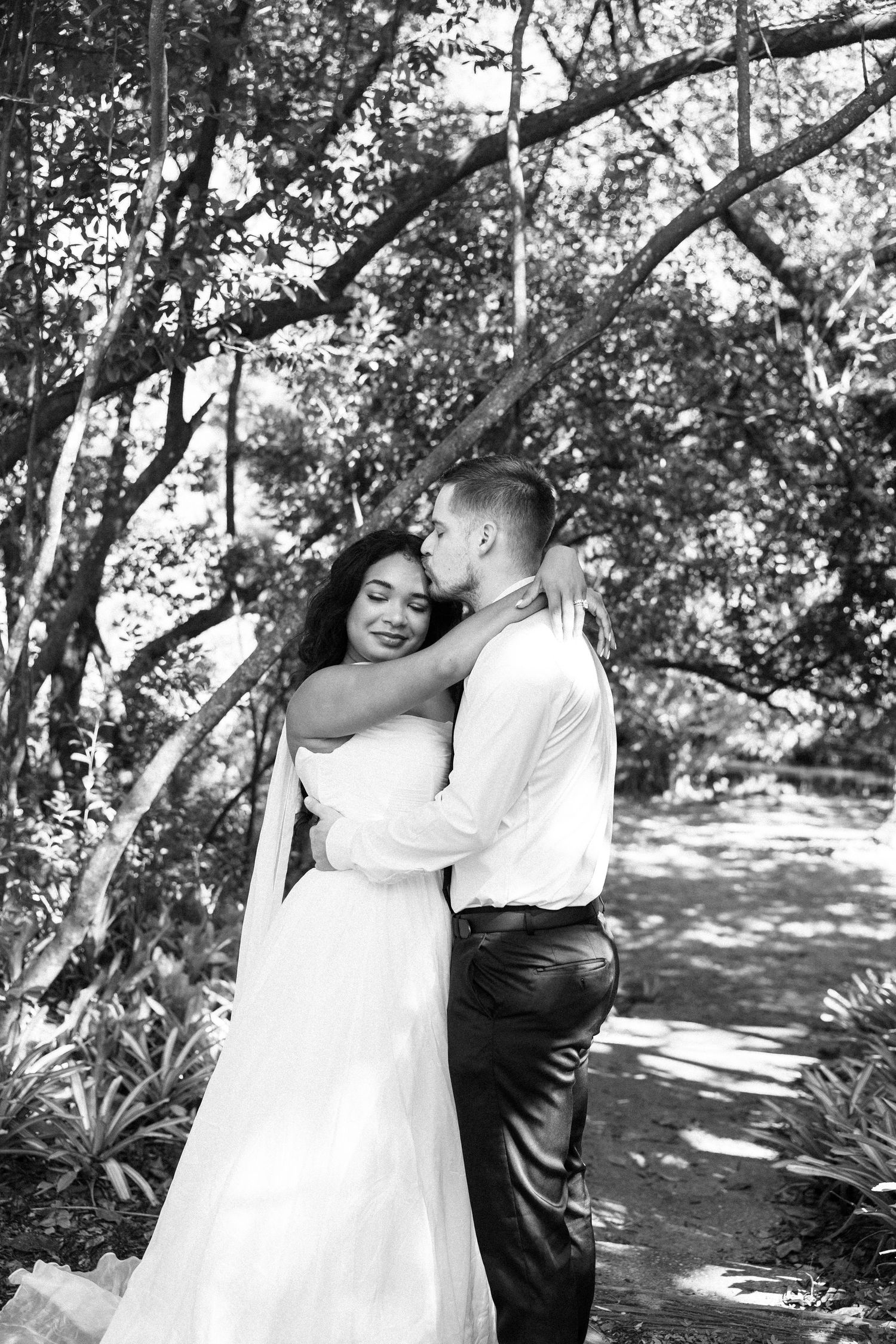 Black and white photo of a bride and groom embracing tenderly among lush trees in an outdoor garden setting.