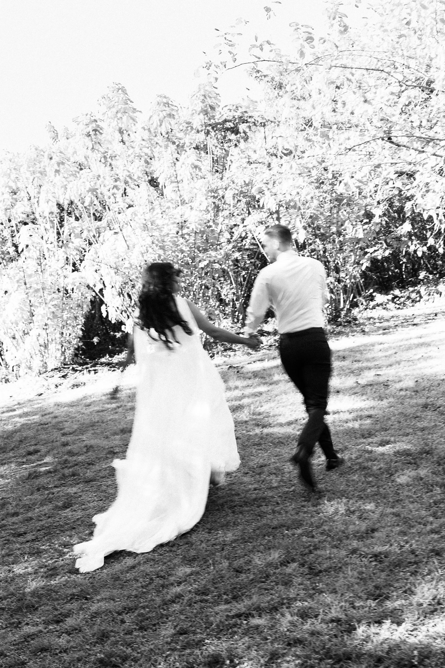 Black and white photo of a bride and groom holding hands, running across a grassy lawn toward trees.