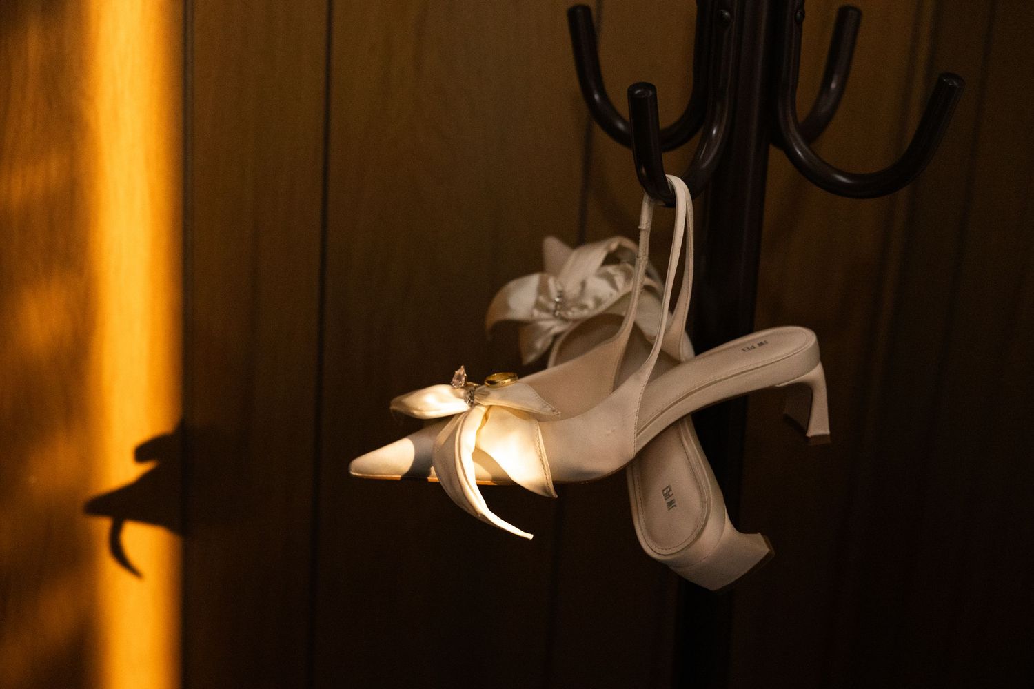 White bridal heels with satin ribbons hanging on a black coat rack in warm golden light.