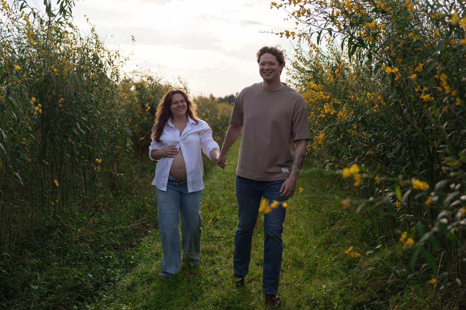 Pregnant woman and man walk hand-in-hand on a lush green path surrounded by wildflowers at dusk.