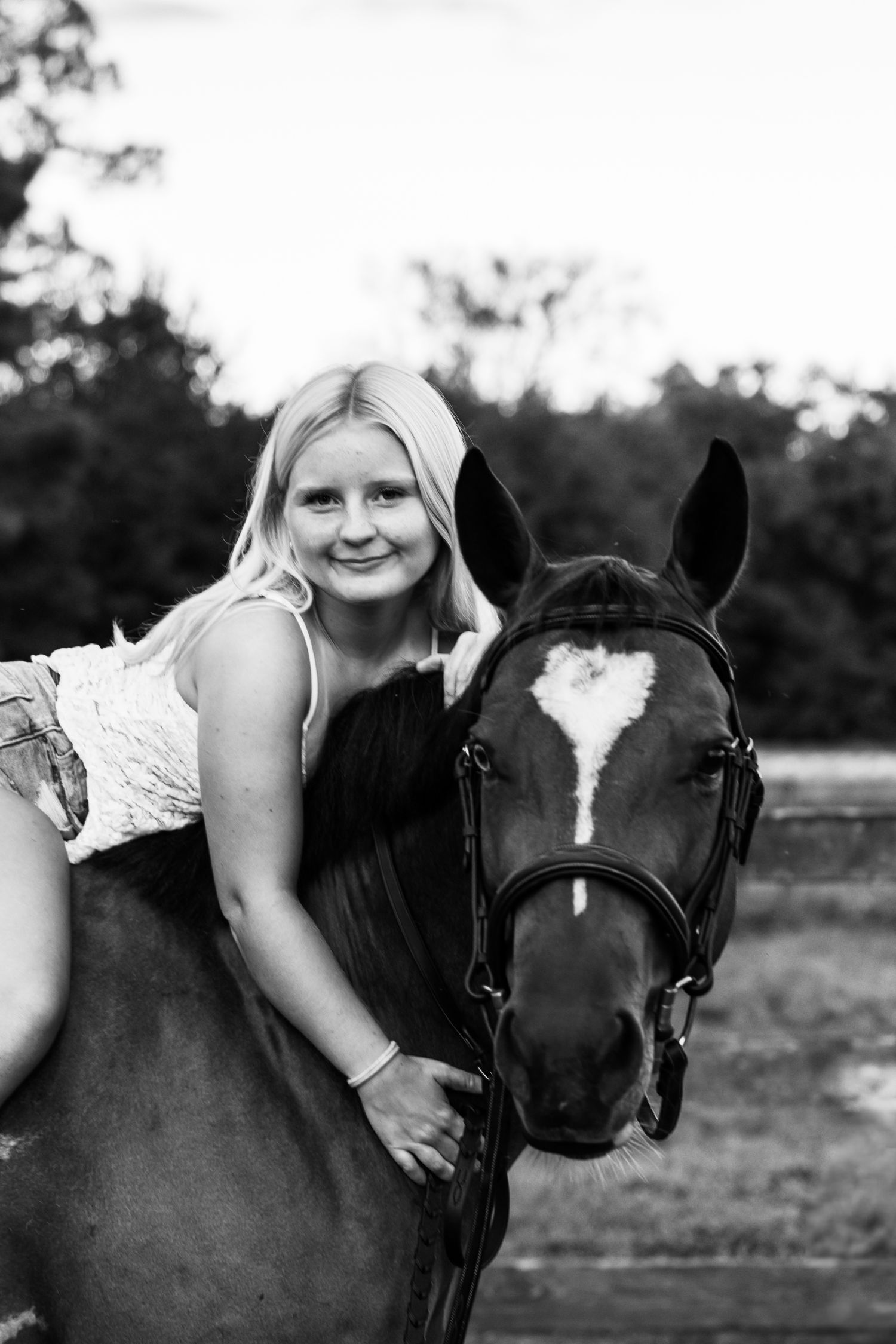 Black and white photo of a smiling blonde girl leaning forward on a dark horse with a white blaze, outdoors.