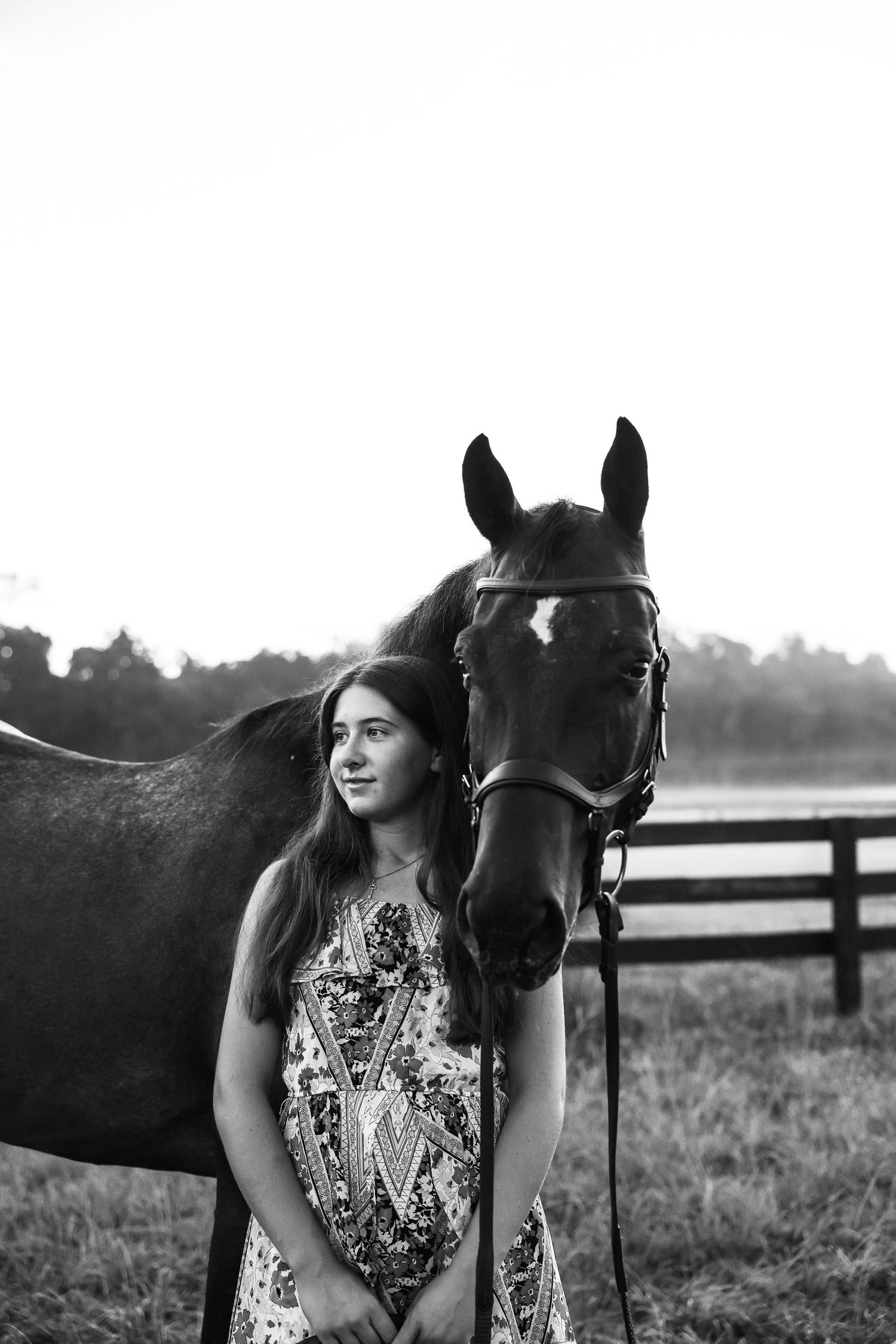 Black and white photo of a young woman in a floral dress standing beside a horse in a fenced pasture.