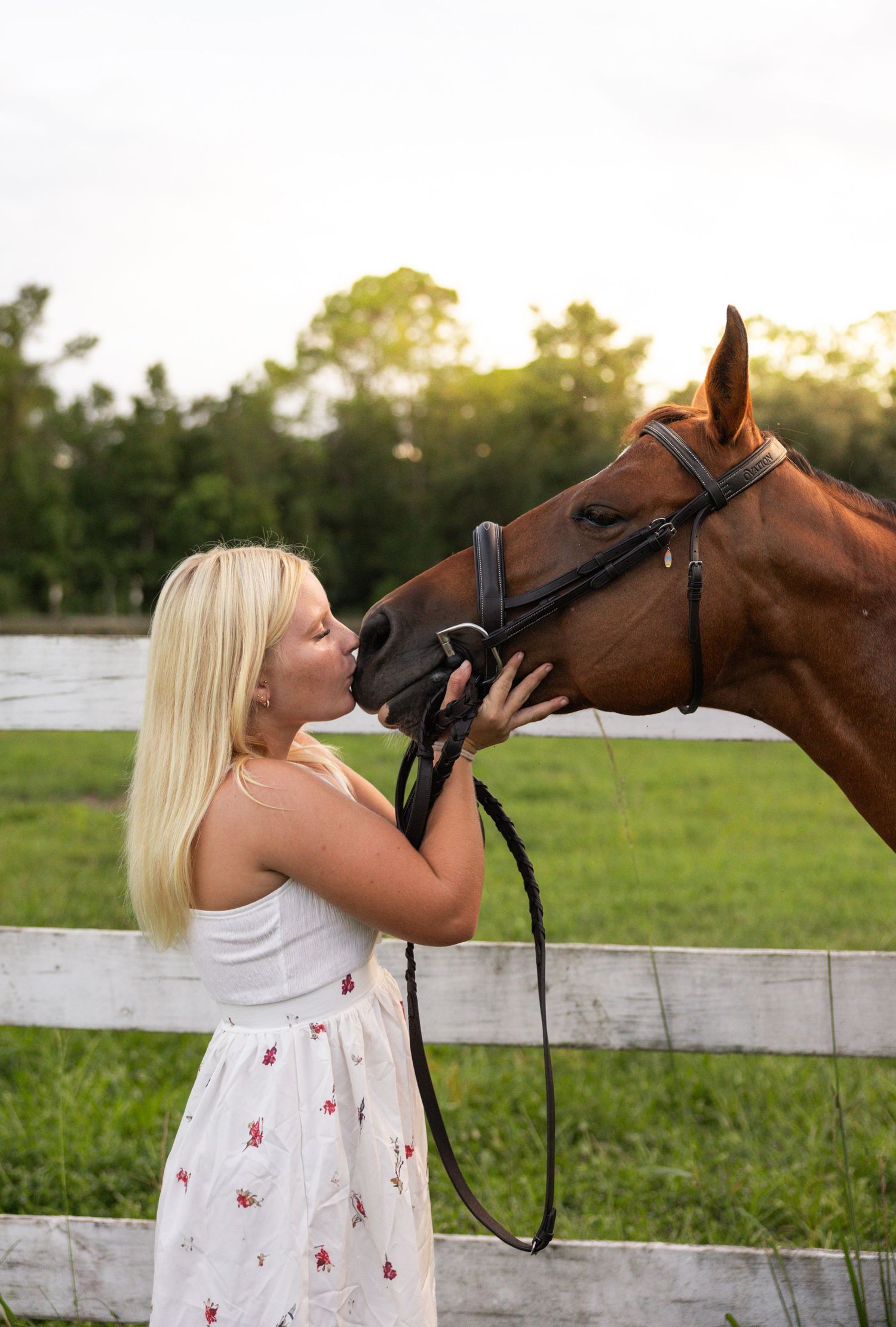 Blonde woman in floral dress kissing a chestnut horse on the nose by a white fence at sunset.