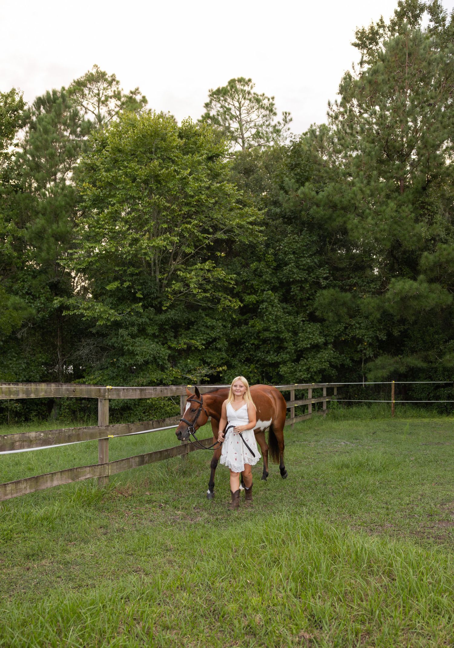 Blonde woman in white dress leading a chestnut horse along a wooden fence in a lush green pasture.