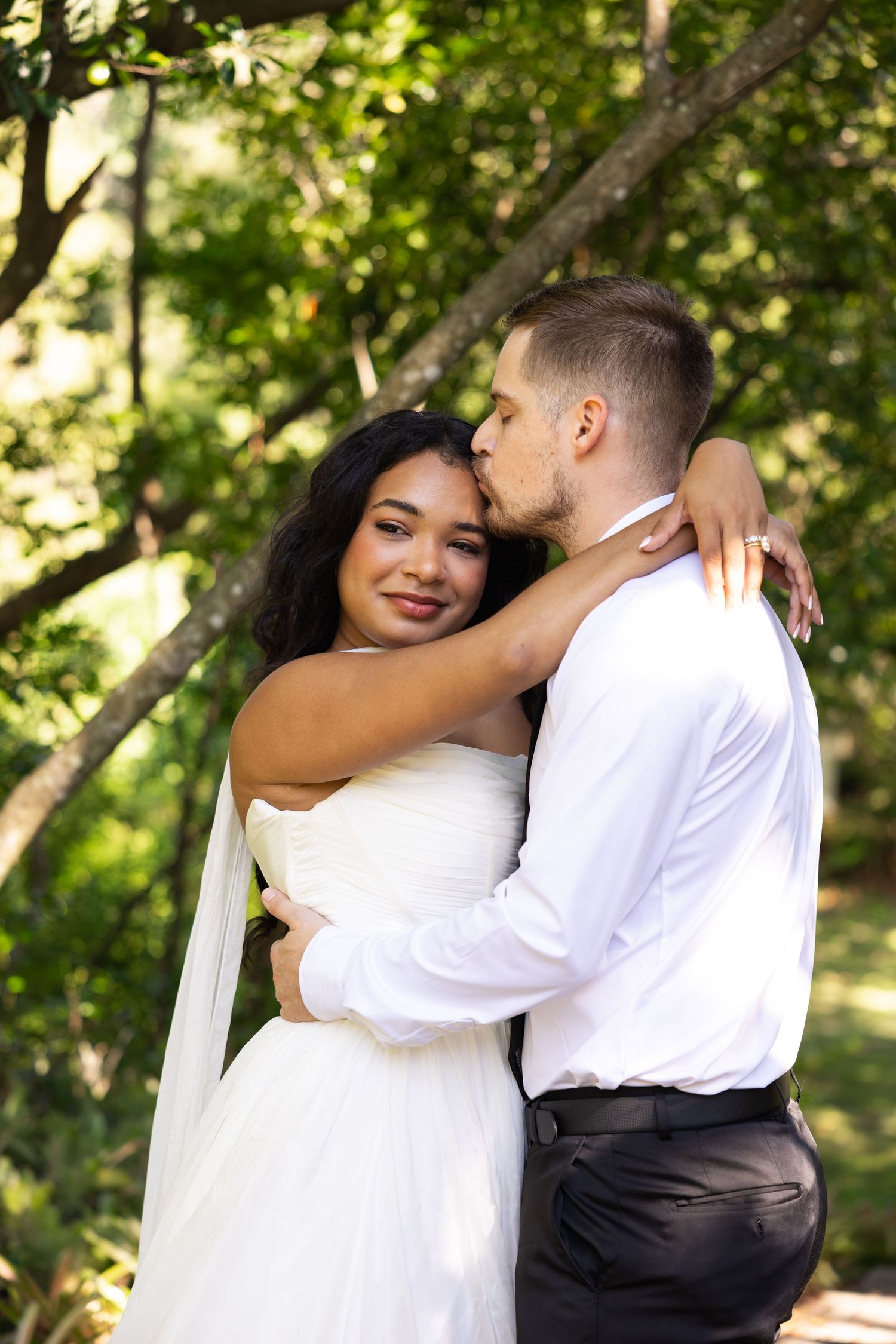 A groom in white shirt kisses his bride in a white gown as they embrace outdoors among lush green trees.