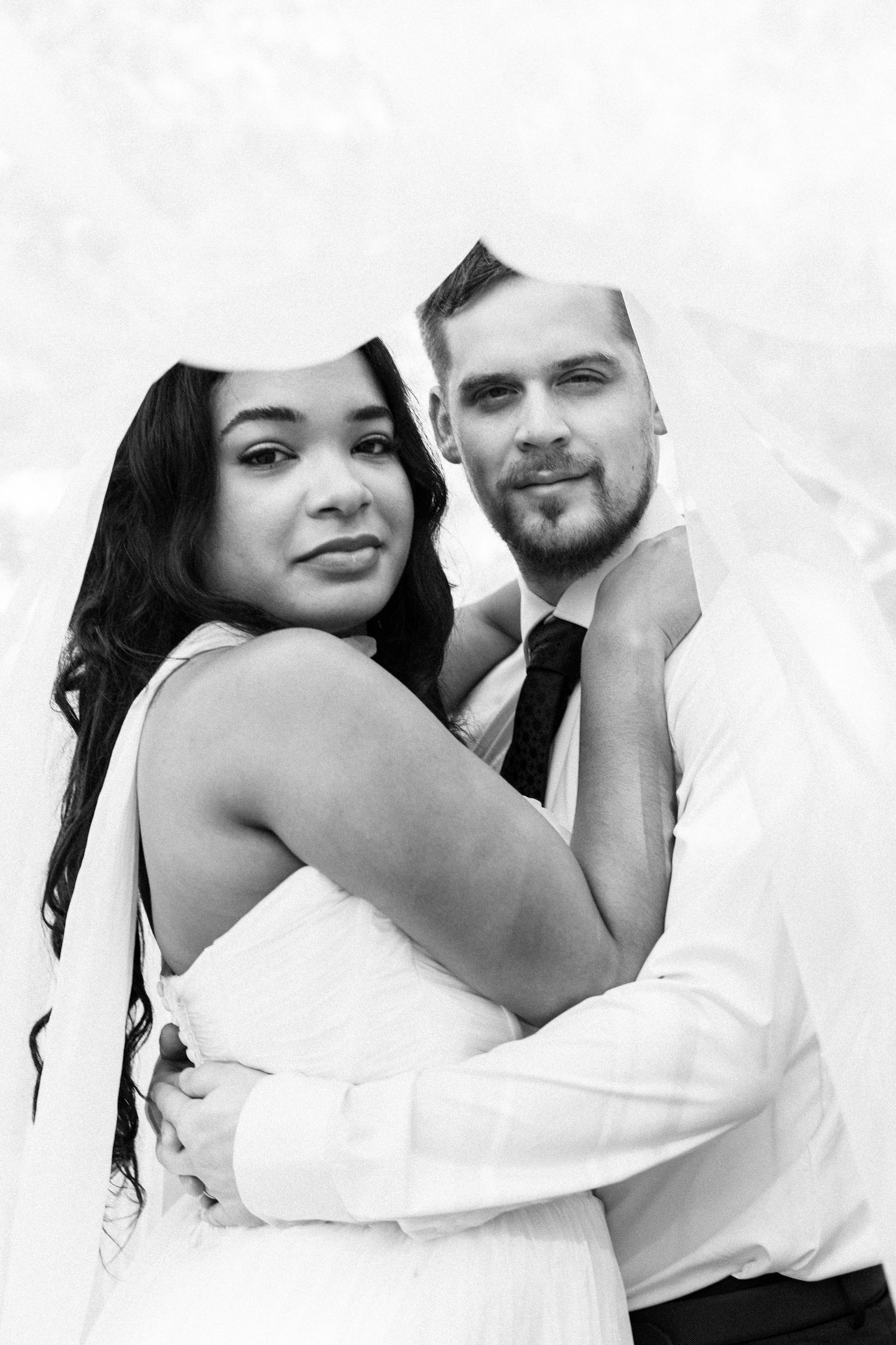 Black and white photo of an interracial couple embracing under a wedding veil, groom in suit, bride in white dress.