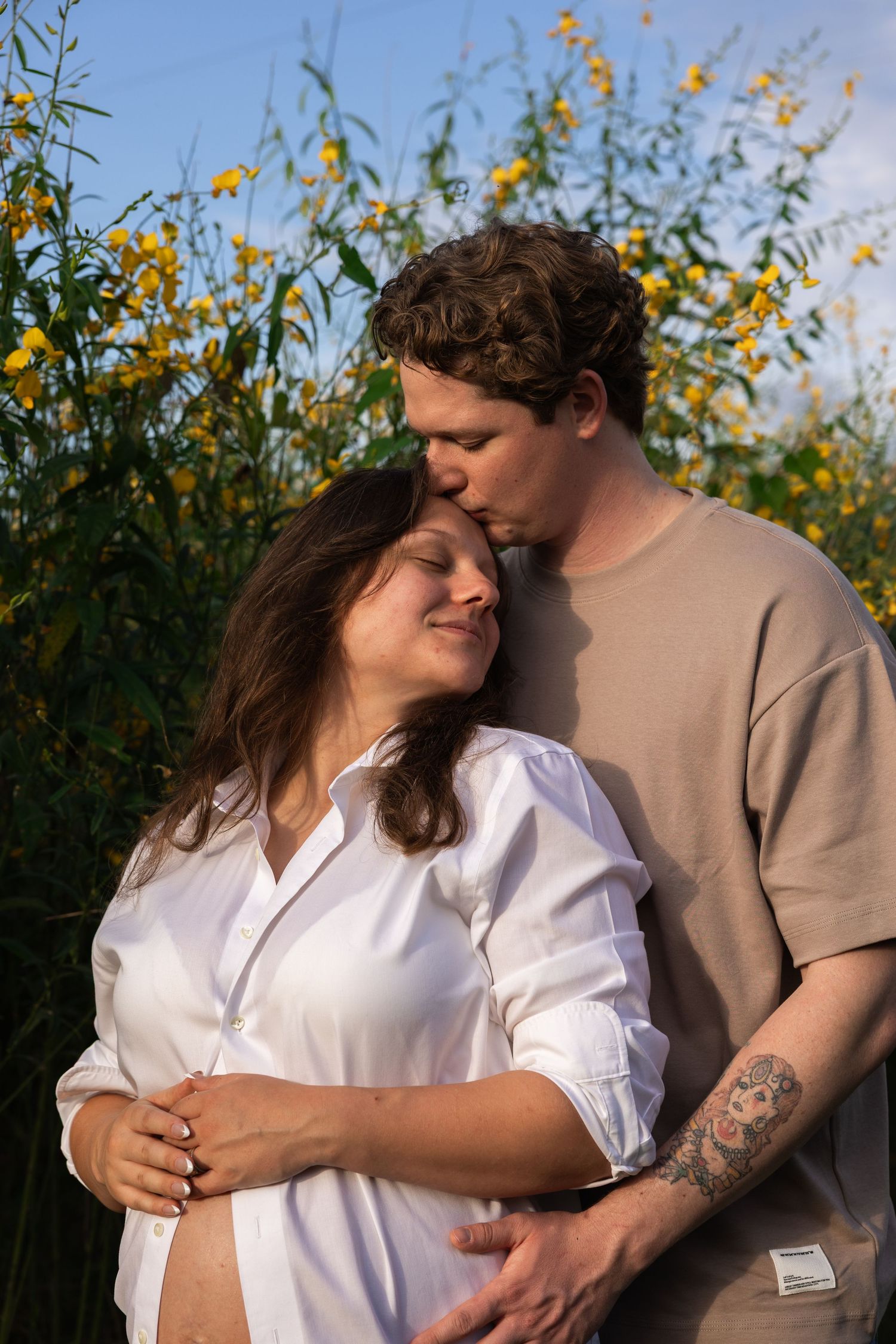 A couple embraces outdoors, the man kissing his pregnant partner's head near yellow wildflowers under a blue sky.