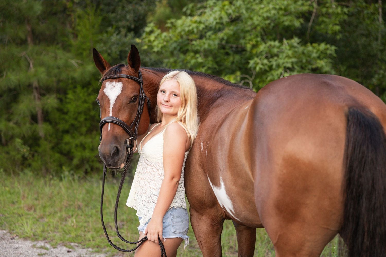 Blonde woman posing with a chestnut horse with a white blaze in a lush green outdoor setting.