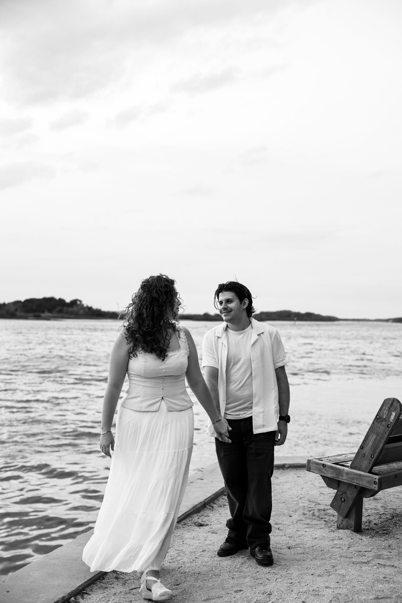 Black and white photo of a couple holding hands on a sandy dock by the water, woman in white dress, man in casual attire.