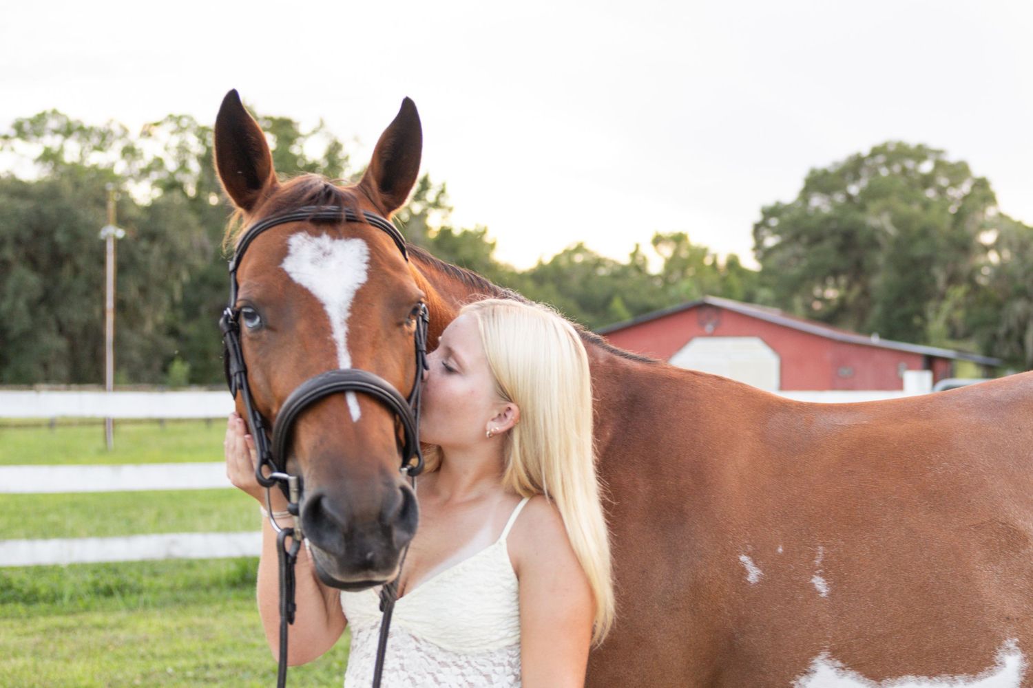 Blonde woman kissing a chestnut paint horse on a sunny farm with green grass and red barn.