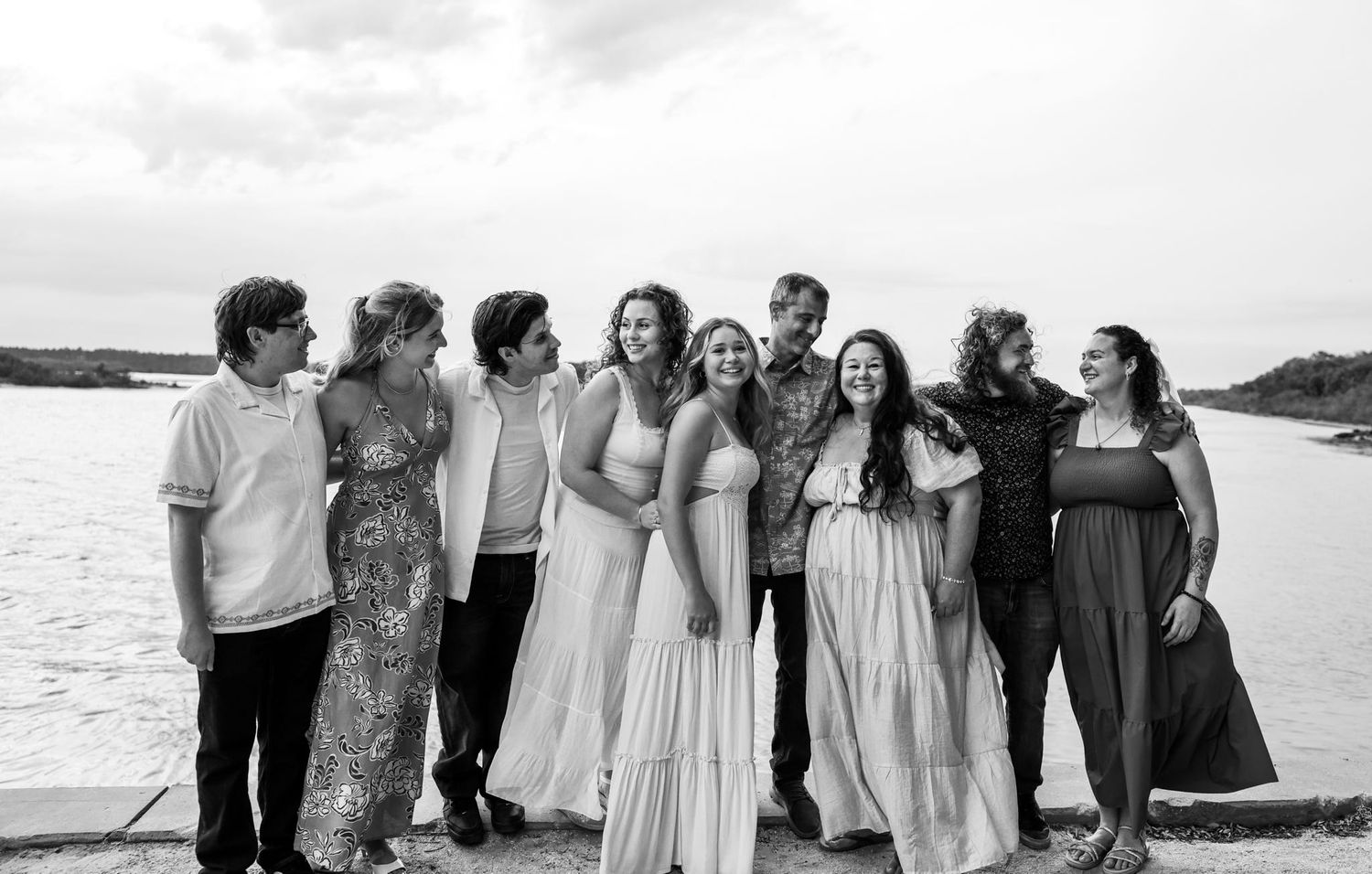 Black and white photo of a large group of friends smiling and laughing together outdoors near a waterfront.
