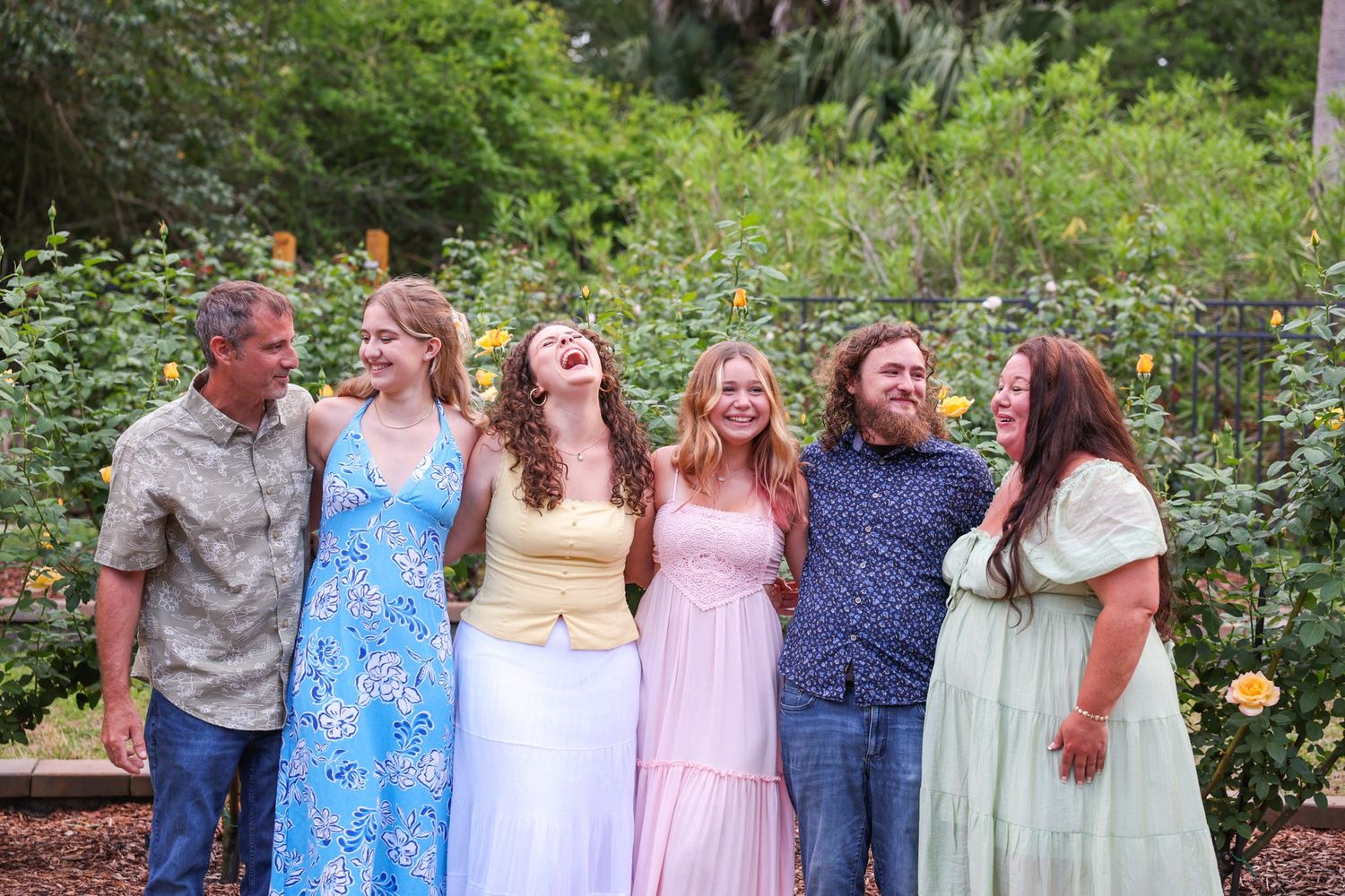 Six people laughing together in a lush garden, wearing colorful summer dresses and casual attire for a family portrait.