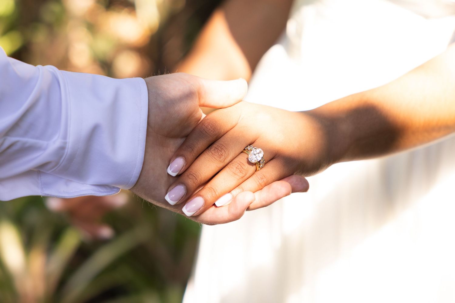 Couple holding hands showing off a diamond engagement ring, bride in white dress and groom in blue shirt outdoors.
