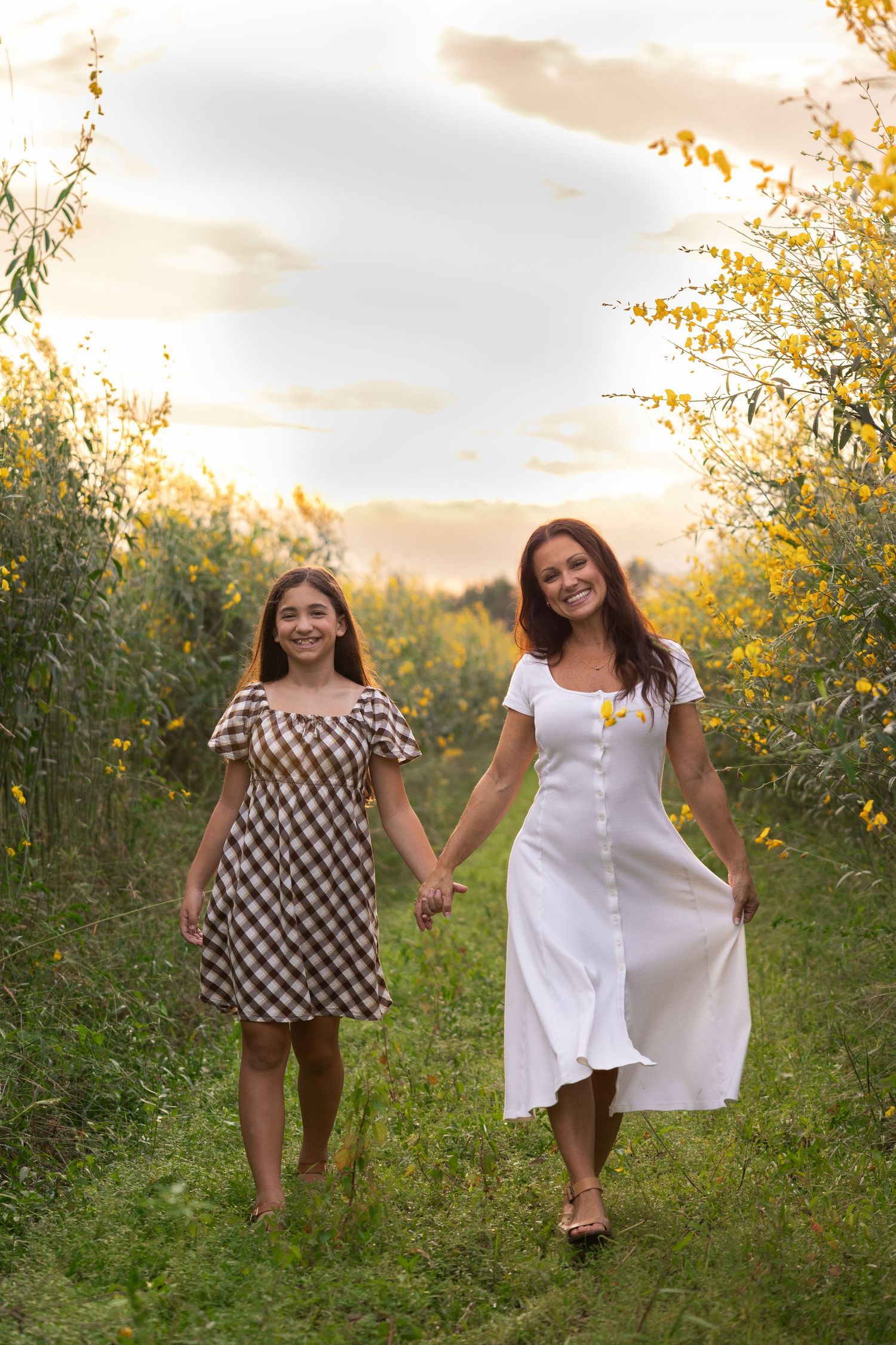 Mother and daughter holding hands walking through a sunny field of yellow wildflowers in summer dresses.