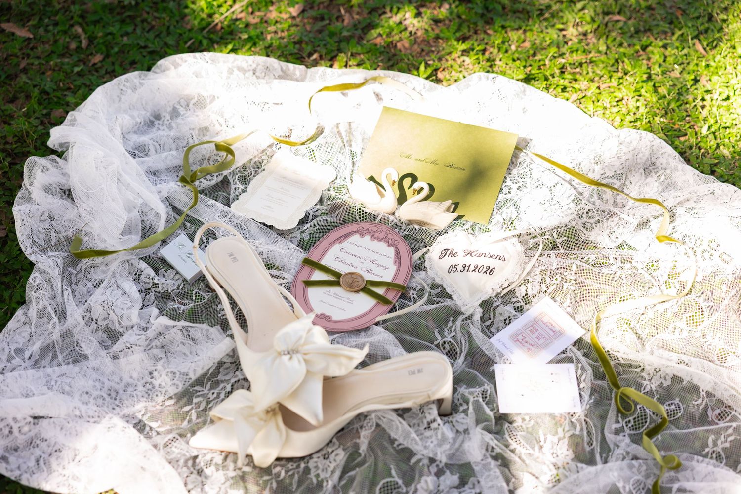 Wedding flat lay with lace veil, heels, pink sunglasses, gold envelope, and handwritten notes on green grass.