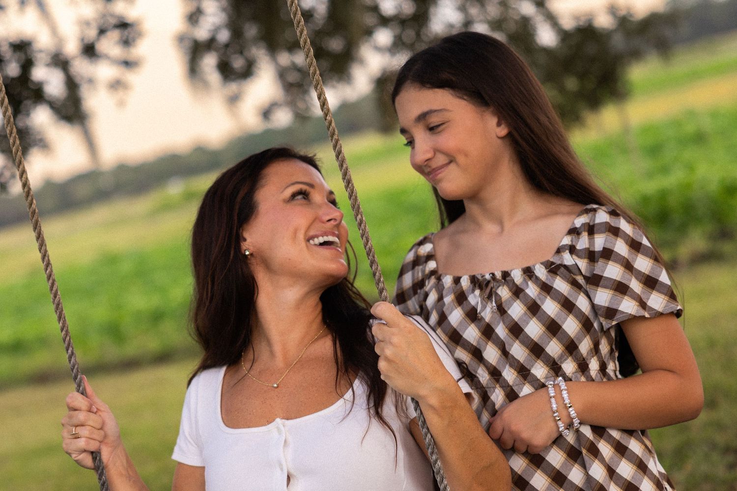 A woman sitting on a rope swing smiles up at a teenage girl in a plaid top outdoors in a green field.