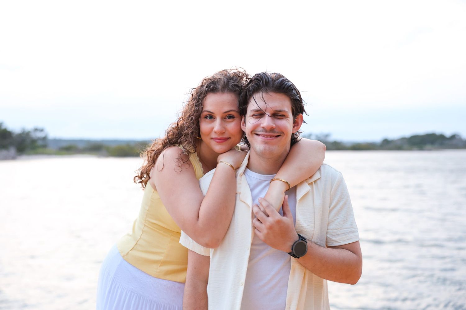A young couple embracing and smiling on a beach at sunset, woman in yellow top hugging man in white shirt.