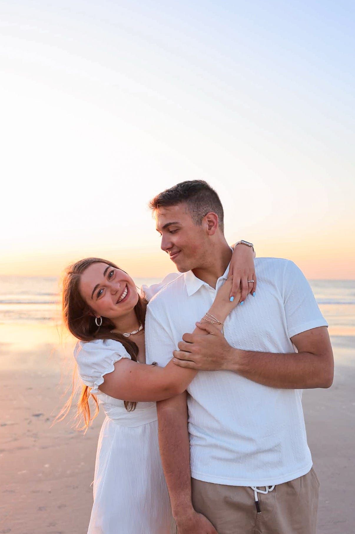 A happy couple embraces on a beach at sunset, both wearing white, sharing a joyful moment by the ocean in North Florida.