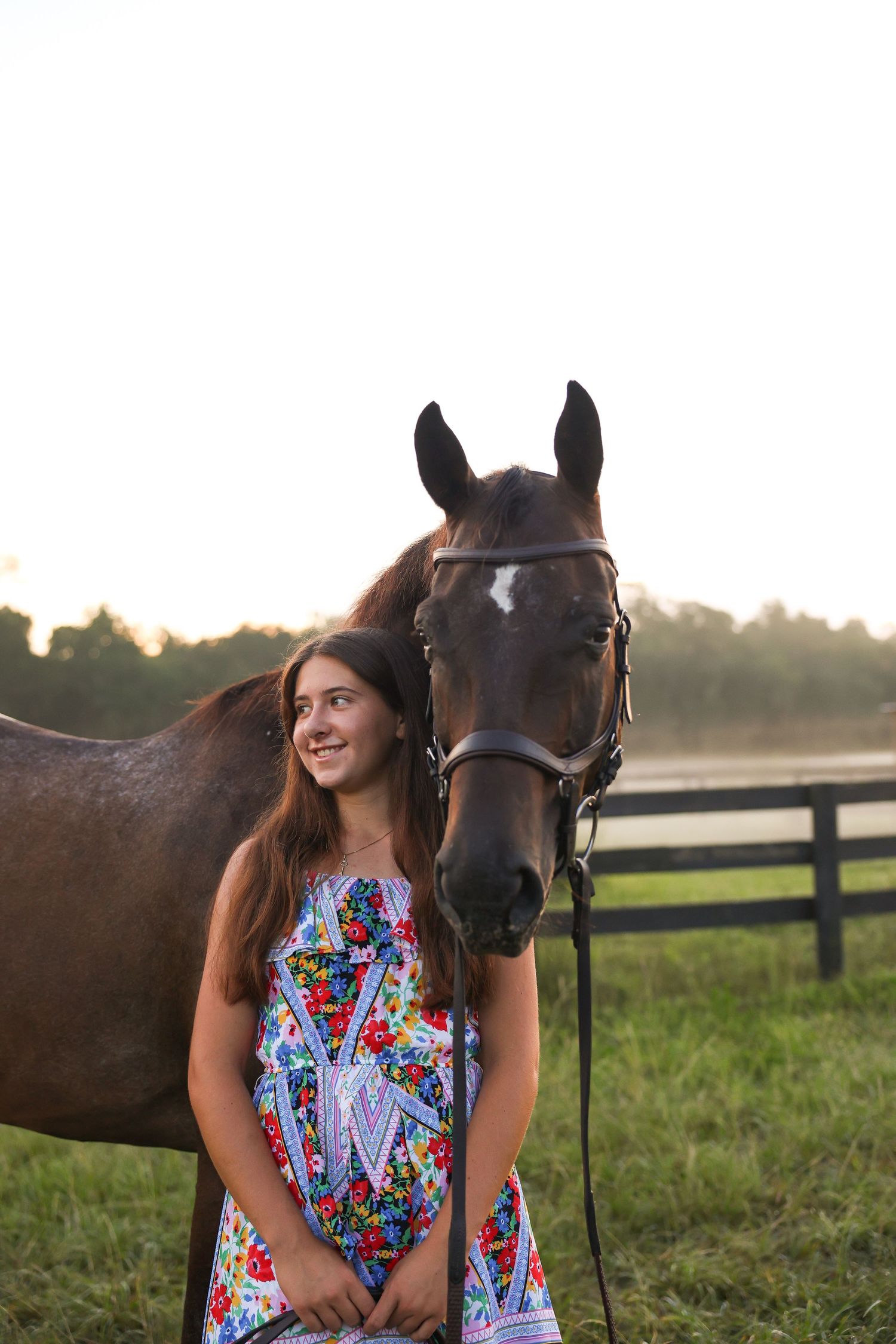 Smiling teenage girl in floral dress standing beside a brown horse at a farm during golden hour sunset.