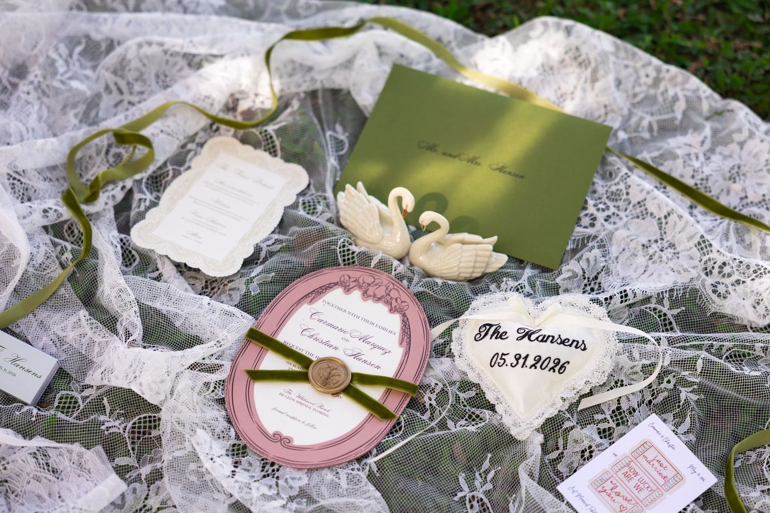 Elegant wedding stationery suite with green envelope, pink circular invite, swan accents, and lace on a garden backdrop.
