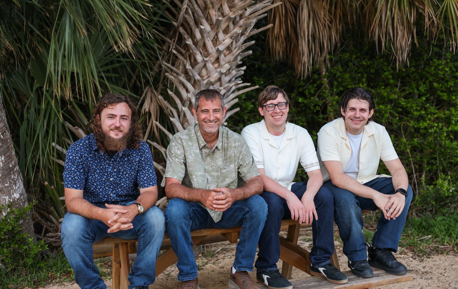 Four men smiling and sitting on a wooden bench outdoors near a palm tree in a casual group photo.