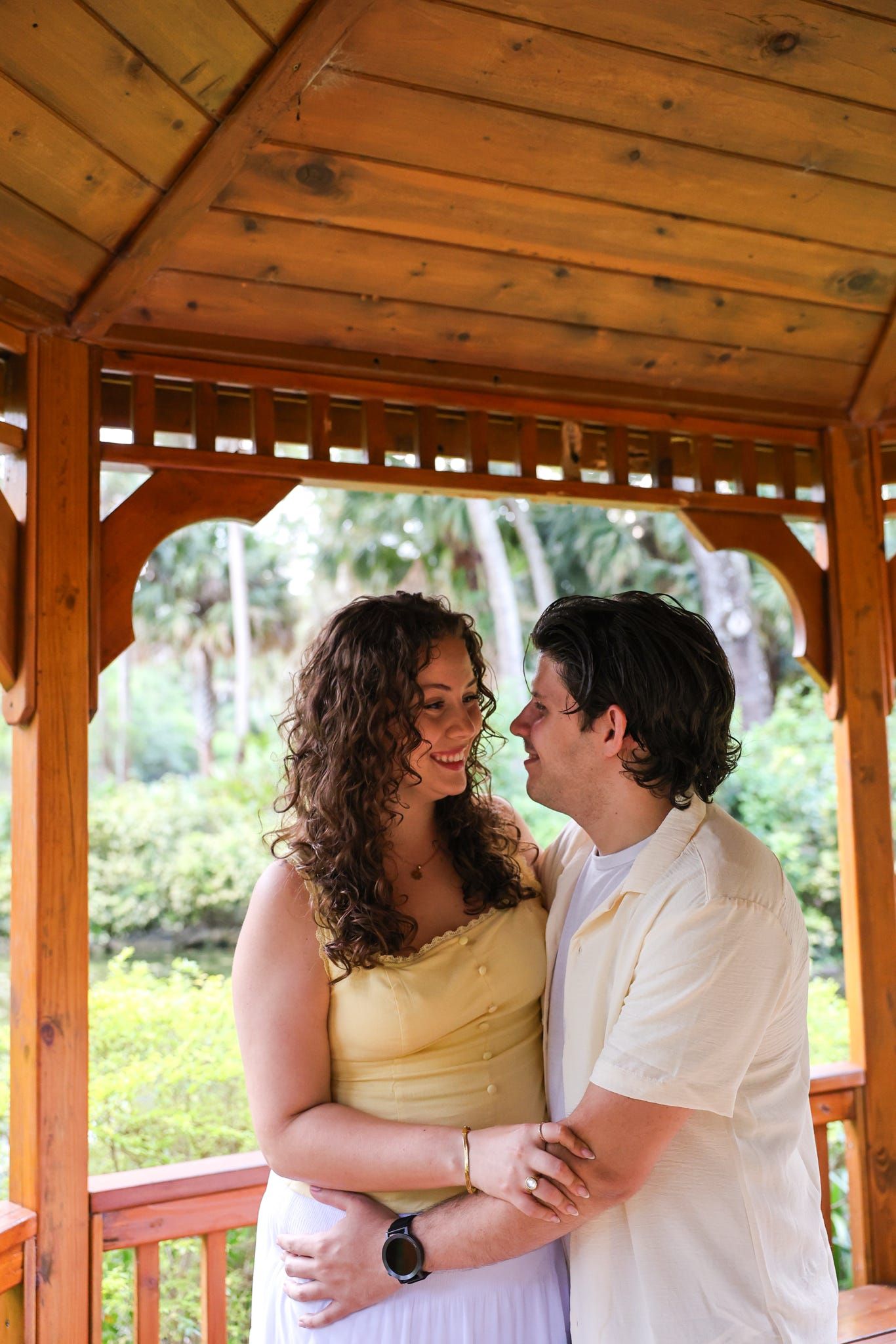 Couple embracing and smiling at each other in a wooden gazebo surrounded by lush green tropical foliage.