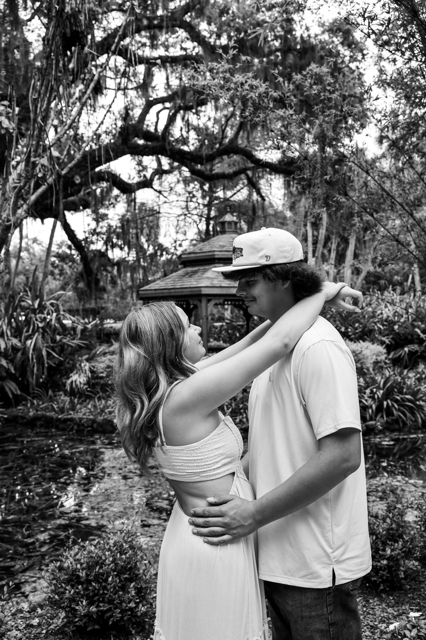 Black and white photo of a romantic couple embracing in a lush garden with moss-covered trees.