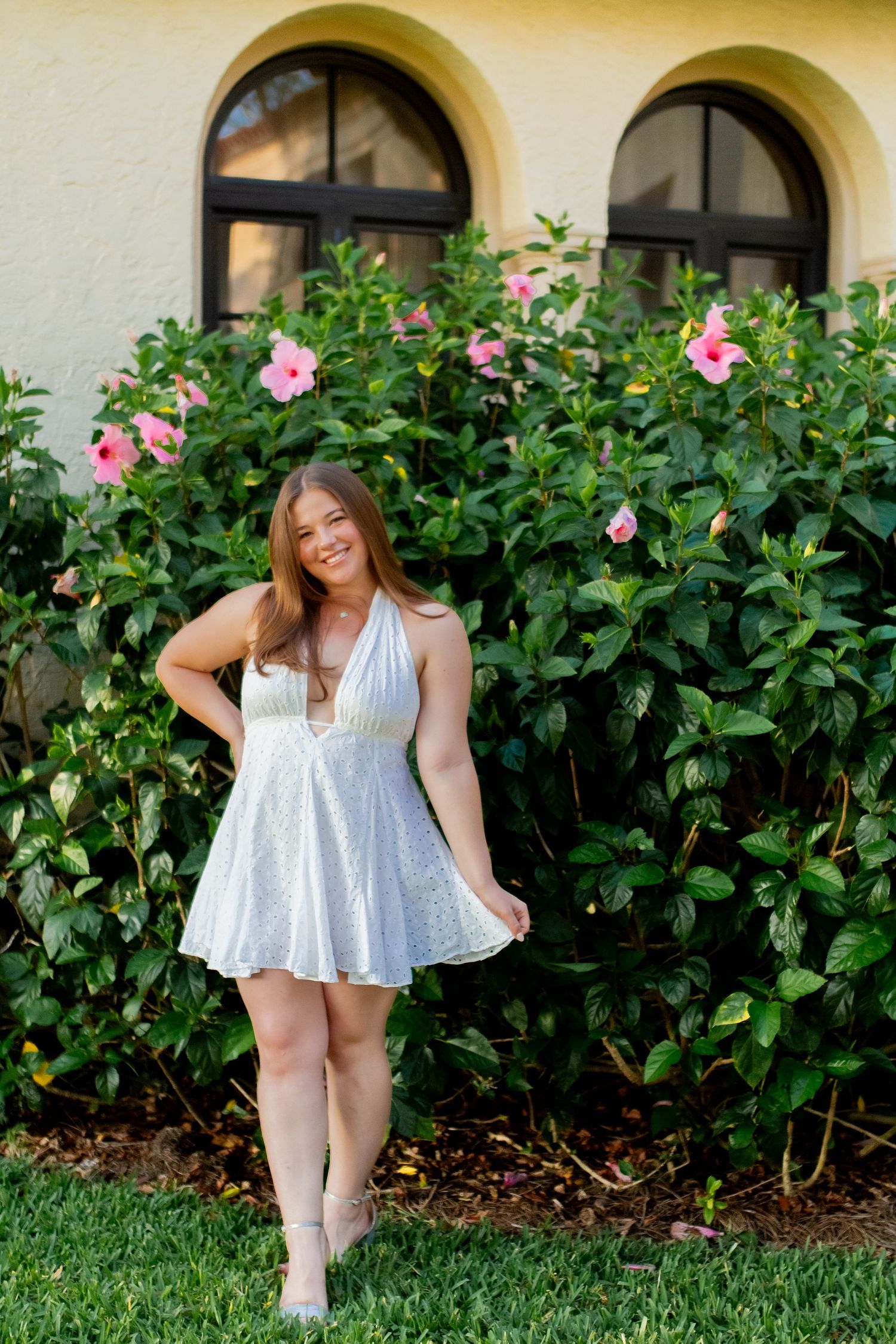 Young woman in white halter dress smiling outdoors near pink hibiscus flowers and arched windows.