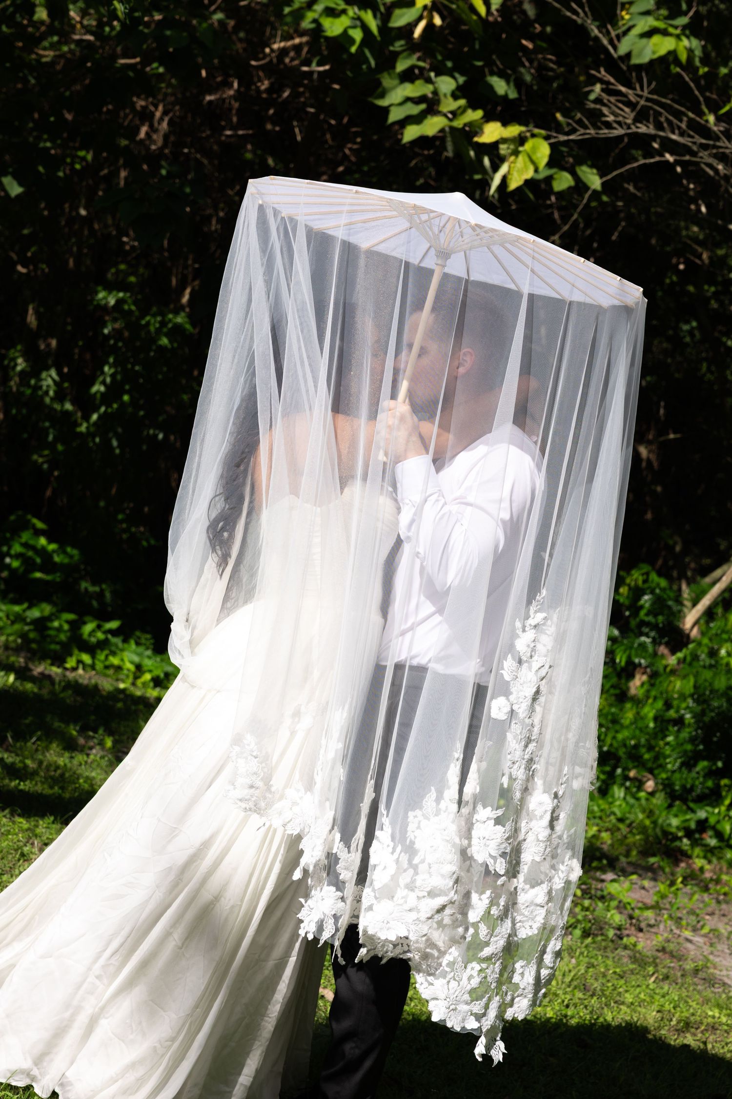 Bride and groom sharing a kiss under a flowing white lace veil outdoors on their wedding day.