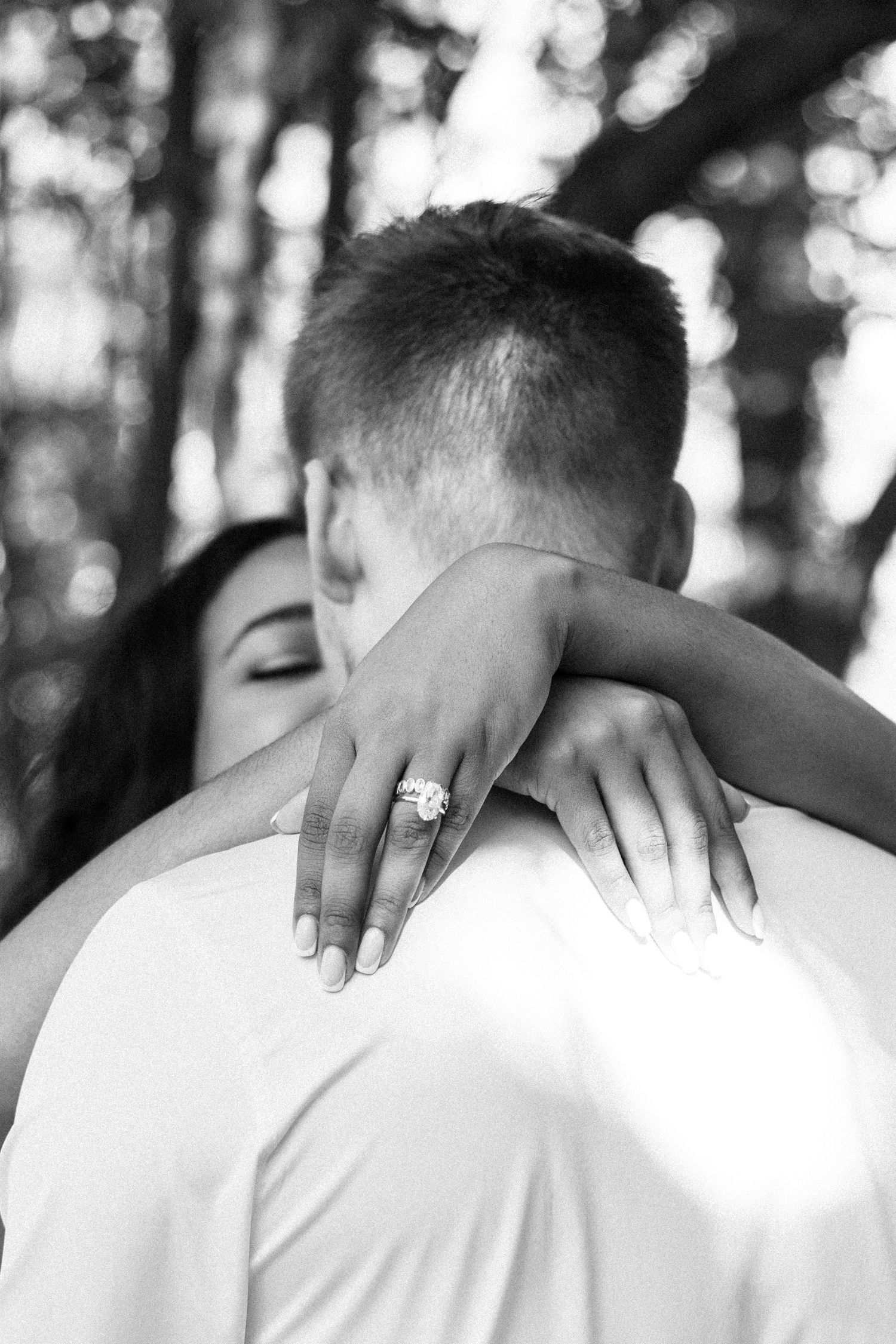 Black and white photo of couple embracing, woman showing diamond engagement ring over man's shoulder.