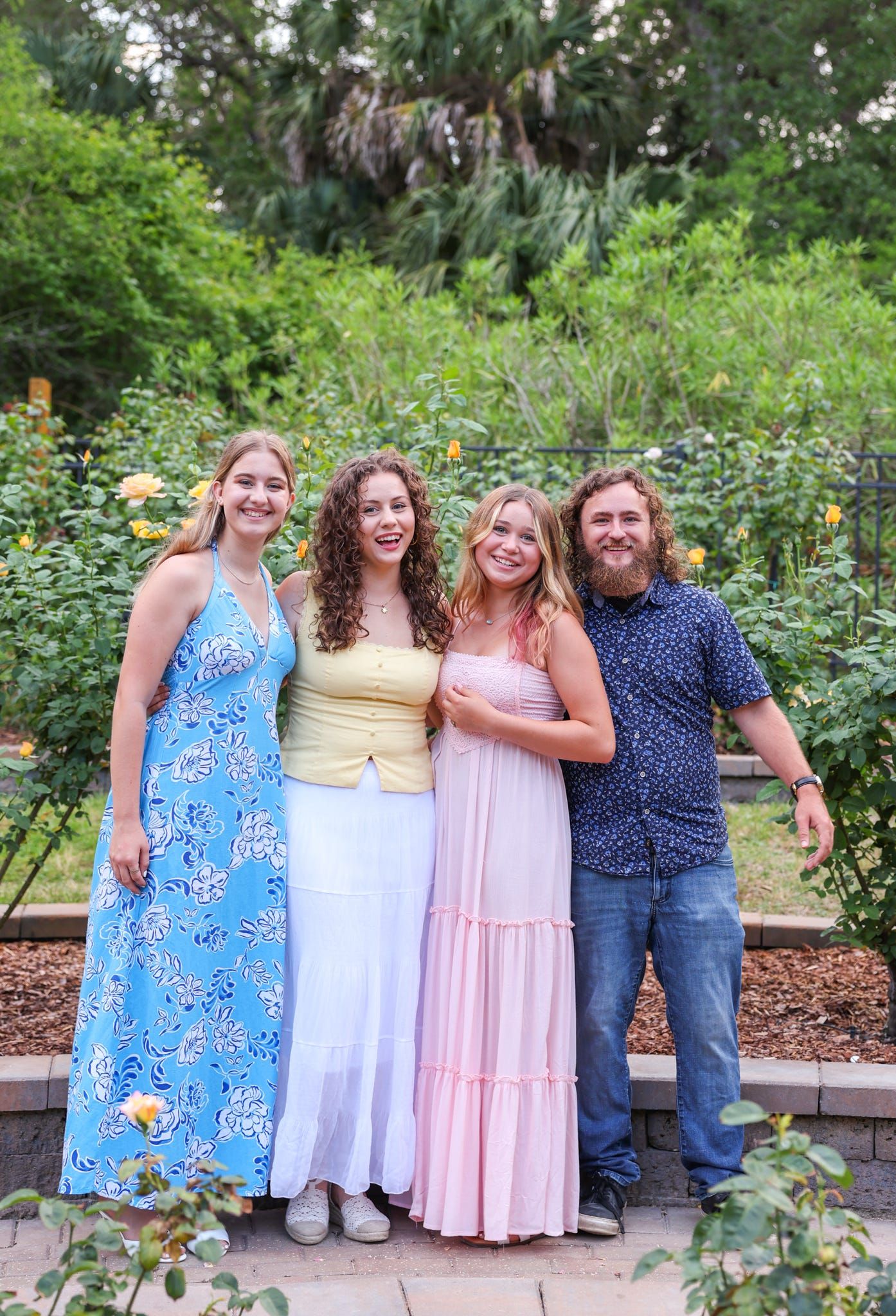 Four smiling people pose together in a lush garden surrounded by yellow roses and green trees on a sunny day.