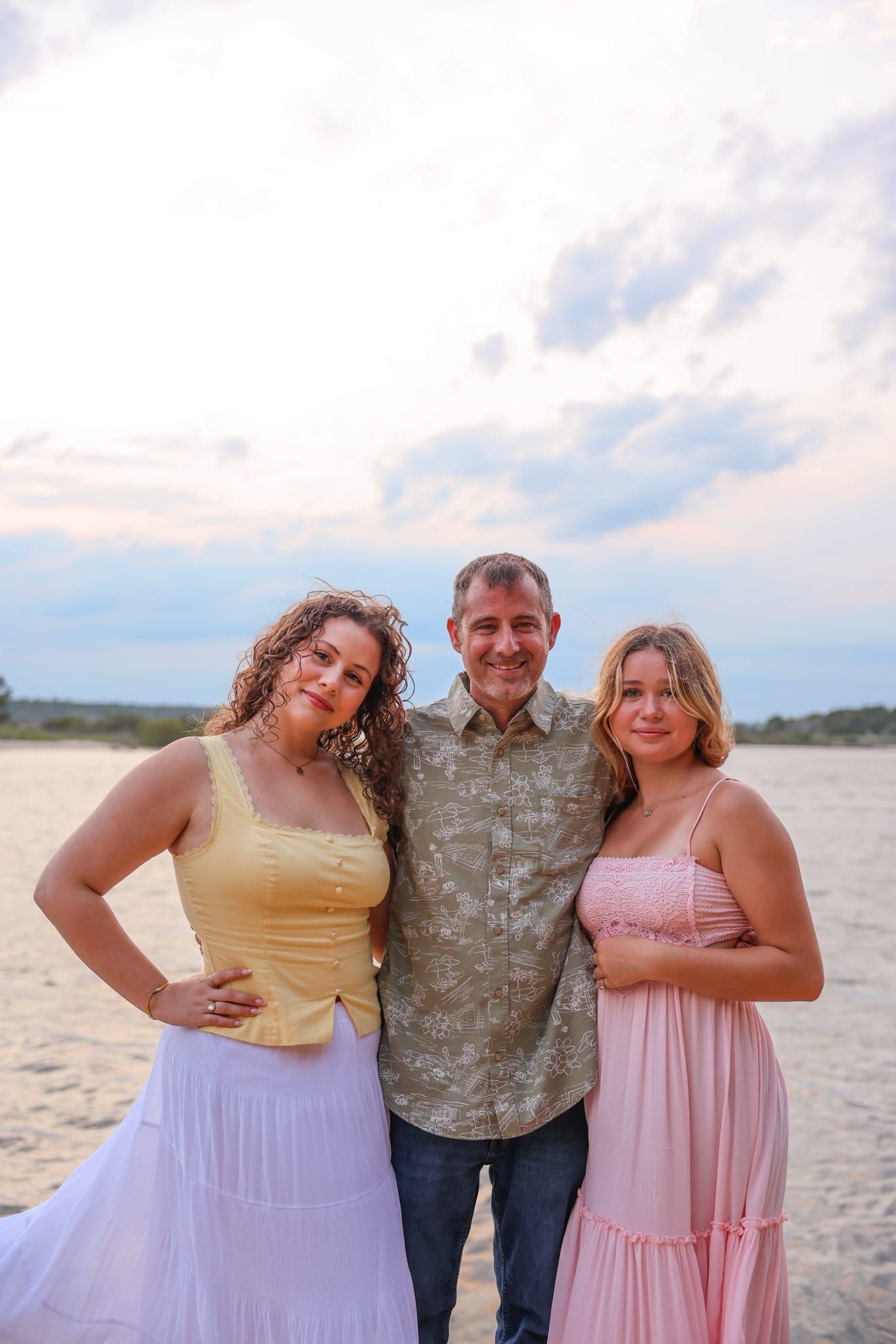 A man and two women smile together on a sandy beach at sunset, wearing summer attire in yellow, floral, and pink.