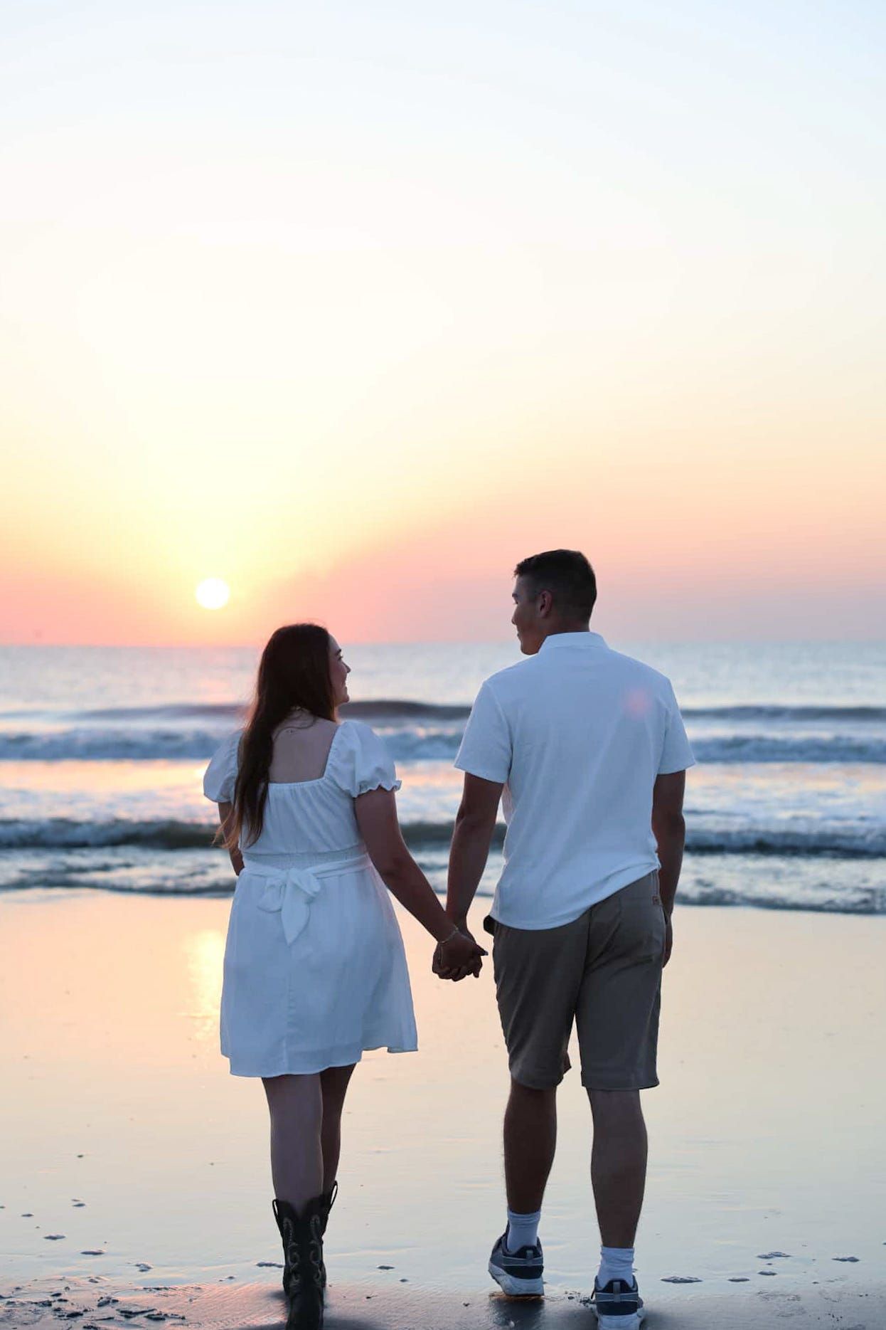 Couple holding hands on beach at sunset, woman in white dress and man in white shirt facing the ocean.