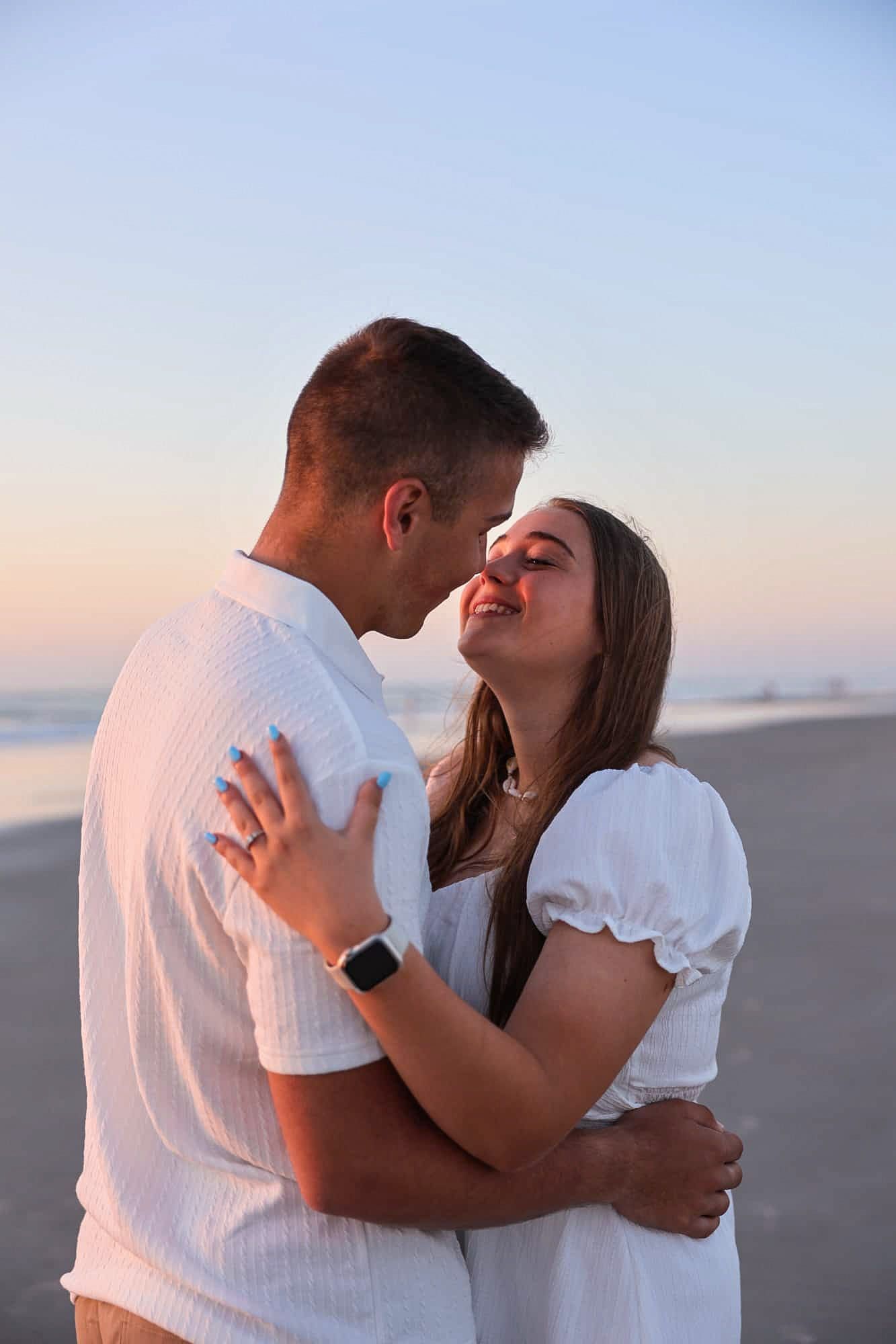 Couple sharing a romantic moment on the beach at sunset, both wearing white outfits near the ocean shore.