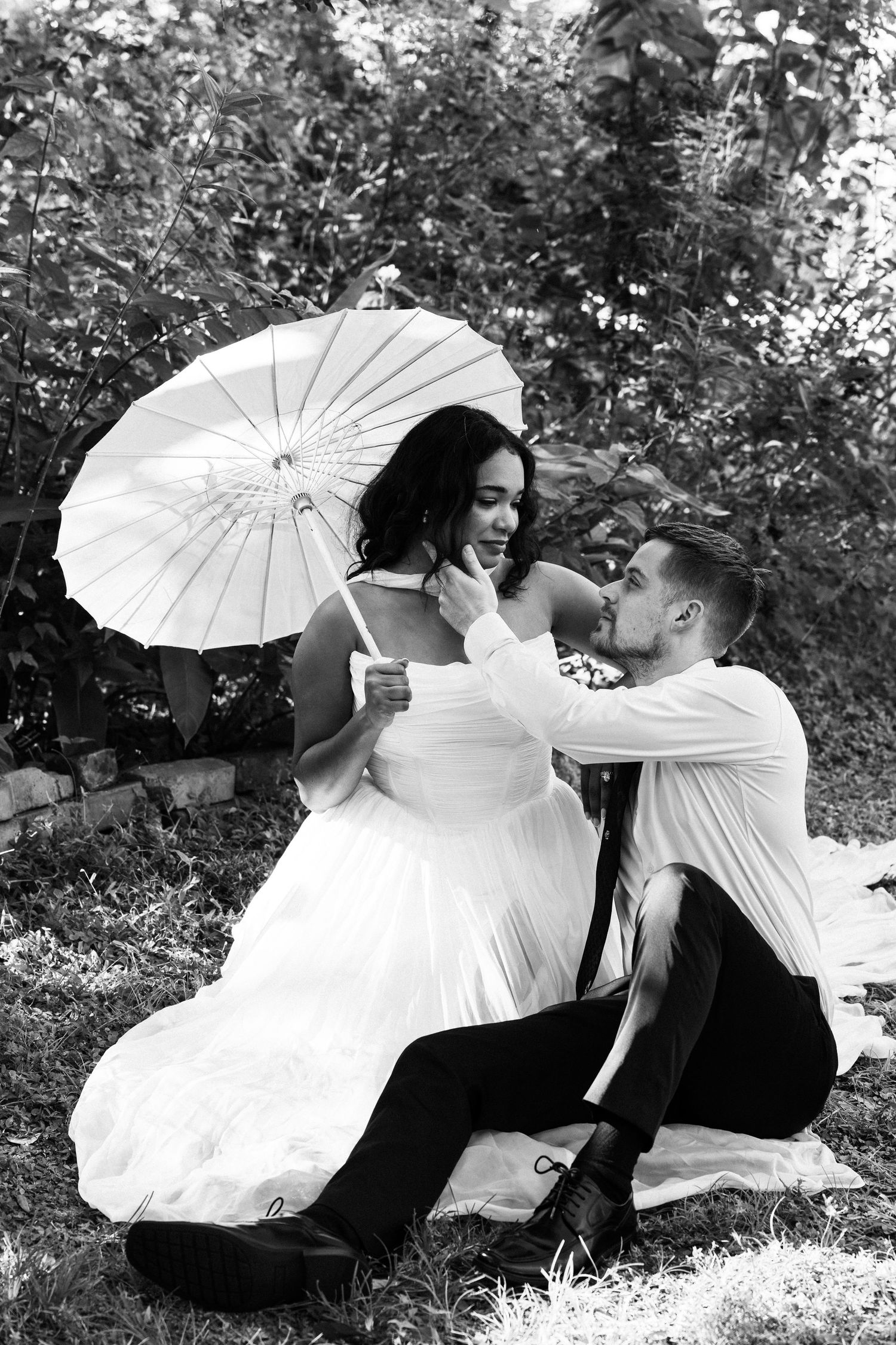 Black and white photo of a groom sitting on grass, tenderly touching brides chin, bride holding white parasol.