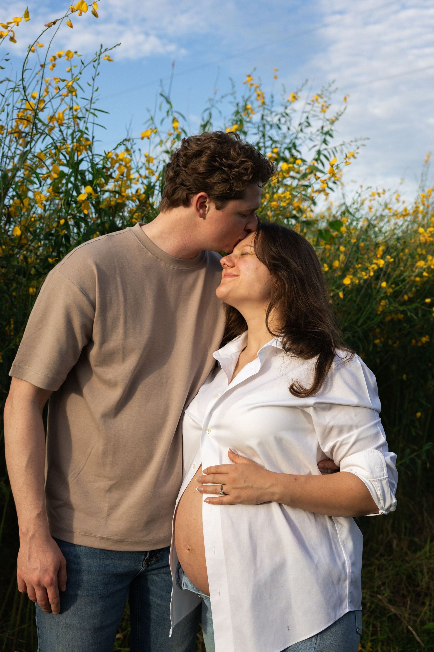 A man tenderly kisses a pregnant woman outdoors, surrounded by yellow wildflowers under a blue sky.