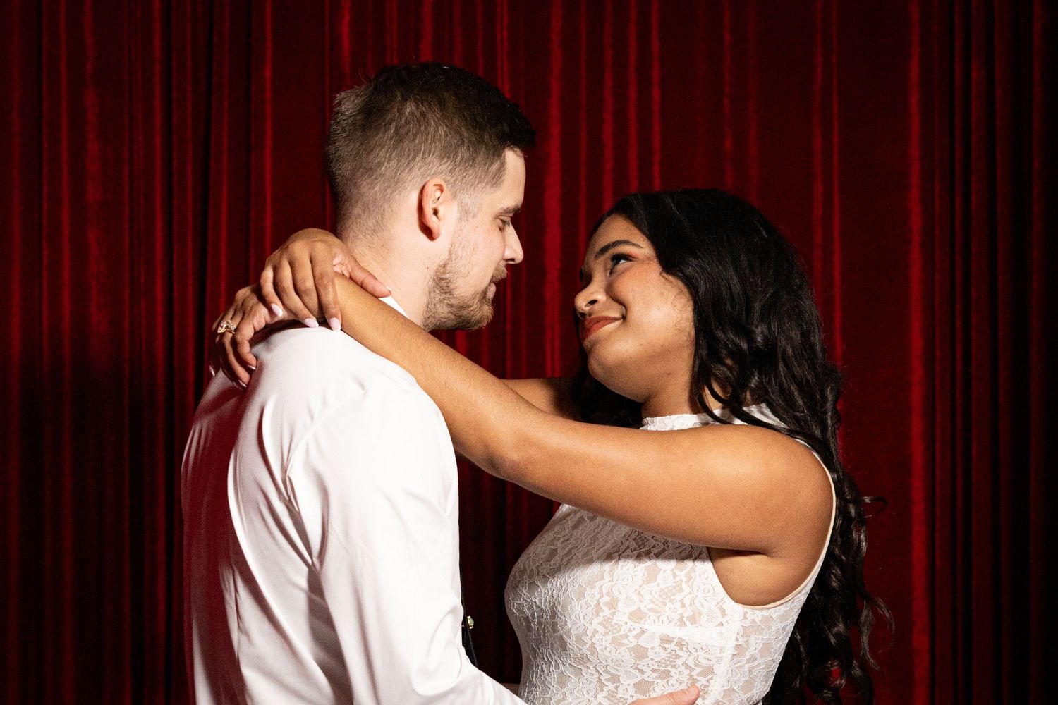 A couple in wedding attire embraces and gazes at each other in front of a red curtain backdrop.