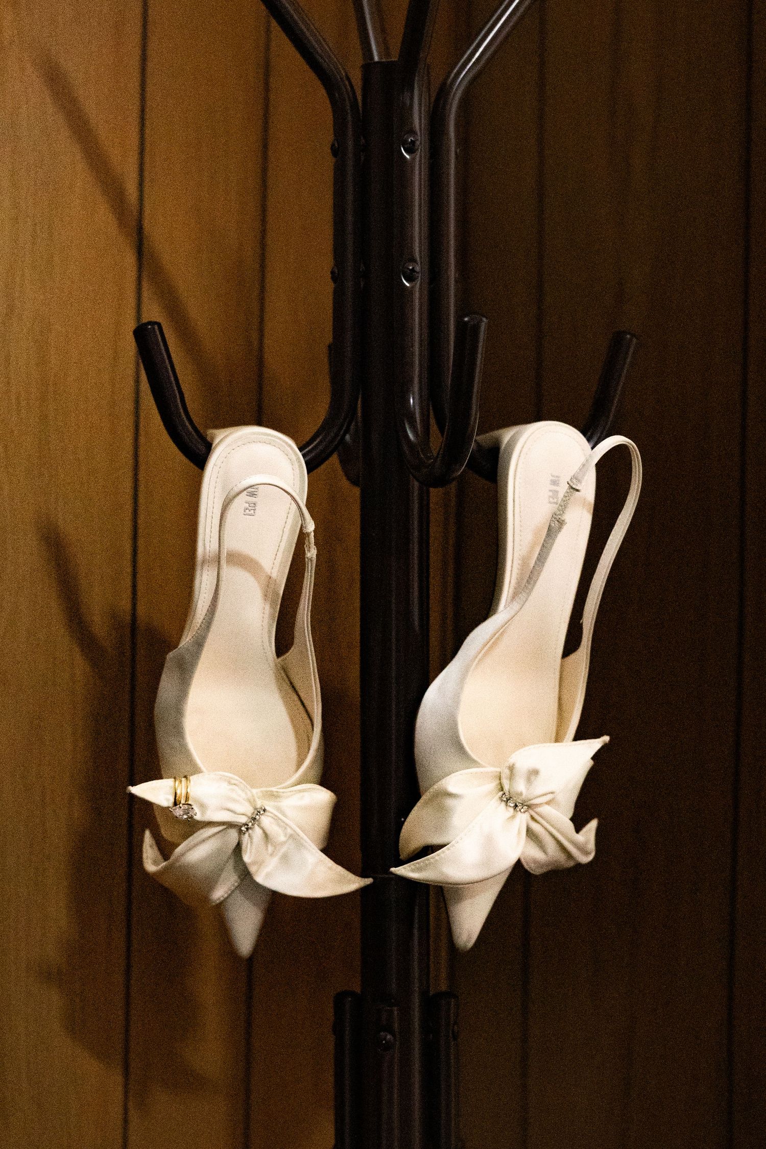 Elegant white satin slingback heels with bow detail hanging on a black coat rack against wooden background.
