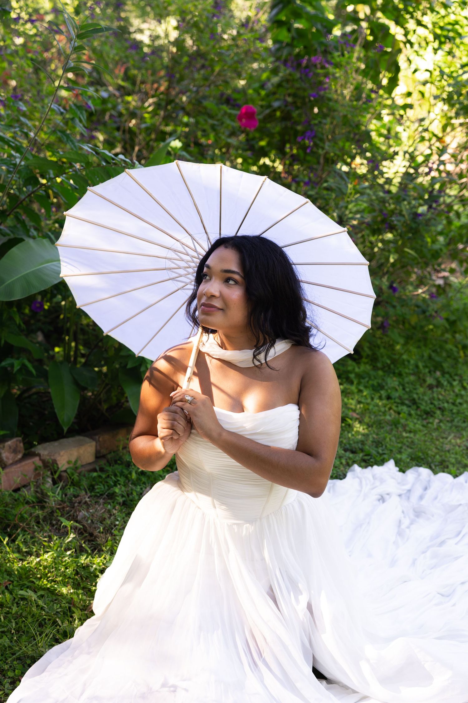 Bride in white strapless gown sits outdoors holding a white parasol umbrella, surrounded by lush green garden foliage.
