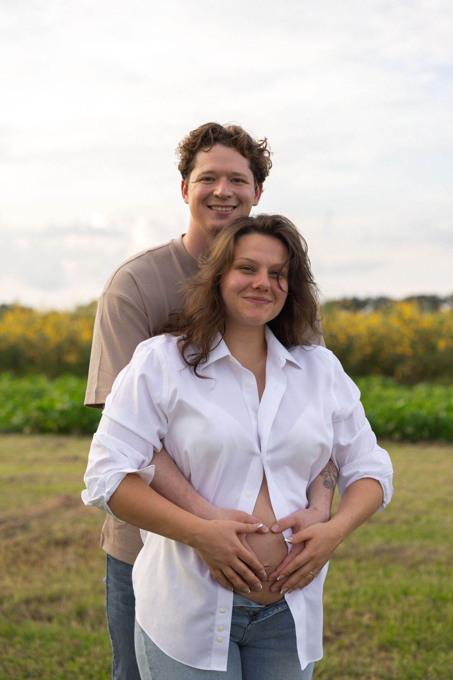 Couple outdoors in field, man embraces pregnant woman from behind, both smiling, forming heart shape with hands on baby bump.