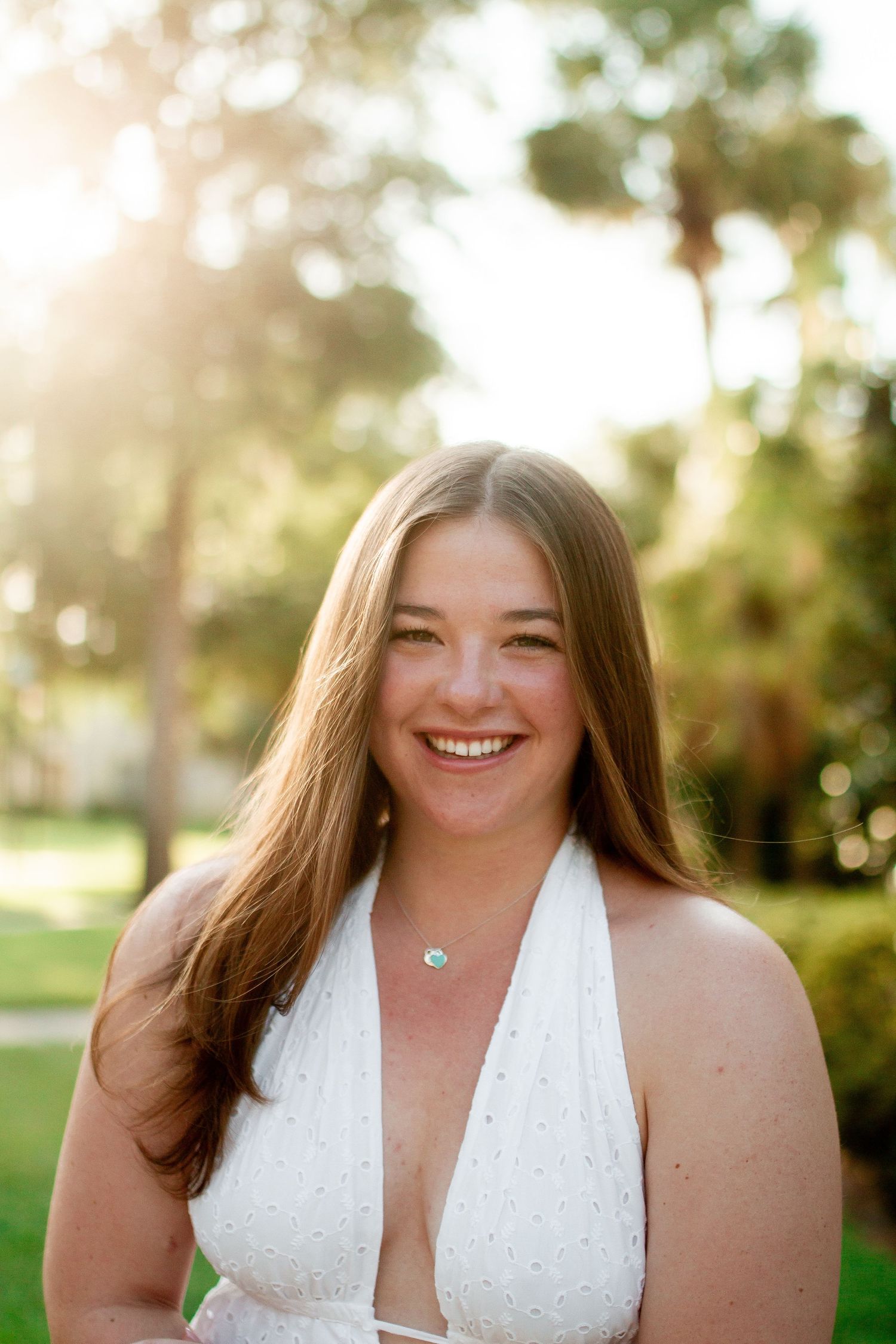 Smiling young woman with long brown hair wearing a white halter dress outdoors with palm trees and sunlight.