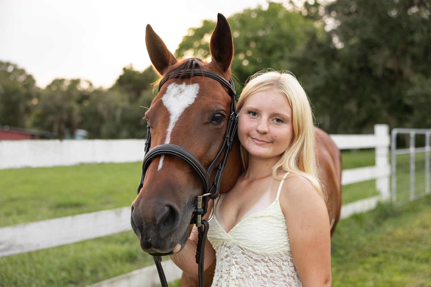 Young blonde woman standing beside a chestnut horse with a heart blaze near a white fence outdoors.