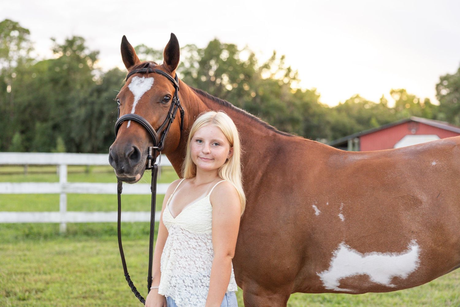 Young blonde girl stands beside a chestnut paint horse in a green pasture with white fence.