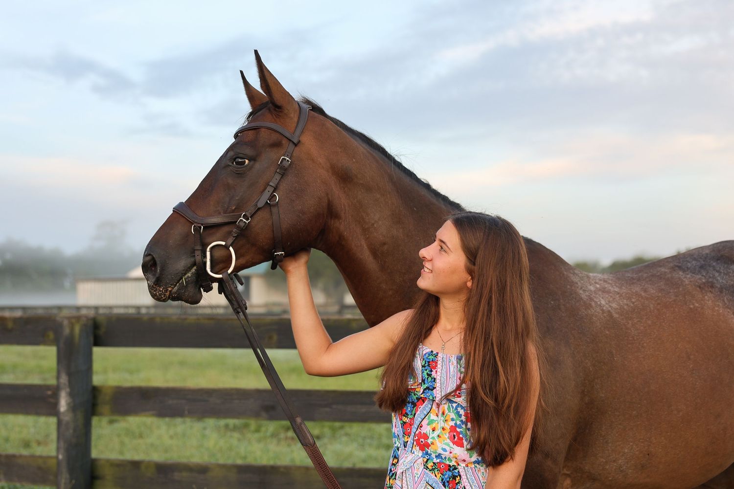 Young woman in floral dress affectionately petting a brown horse outdoors near a wooden fence.