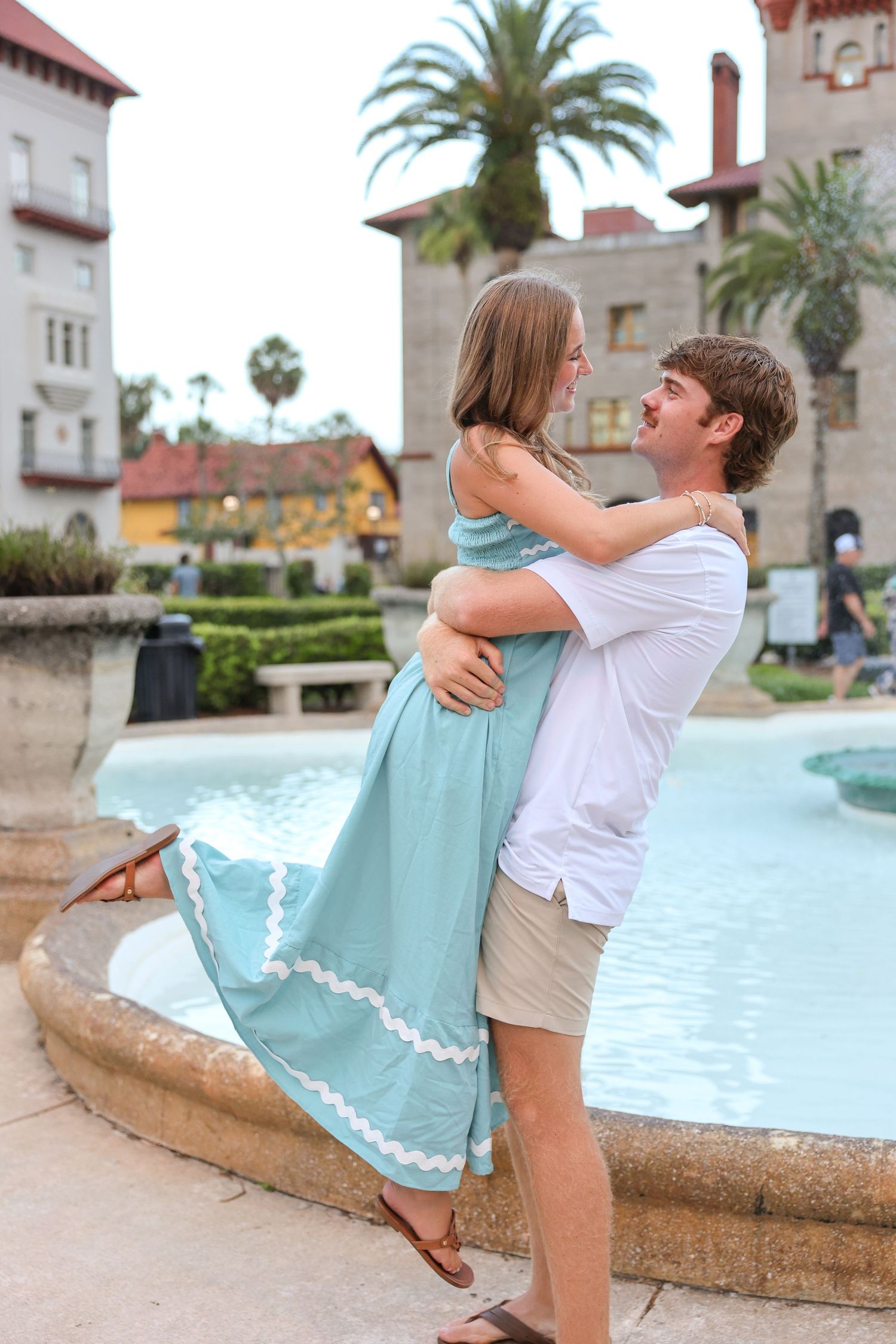 Couple embracing by a fountain, woman in teal dress lifted by man in white shirt, historic building backdrop.