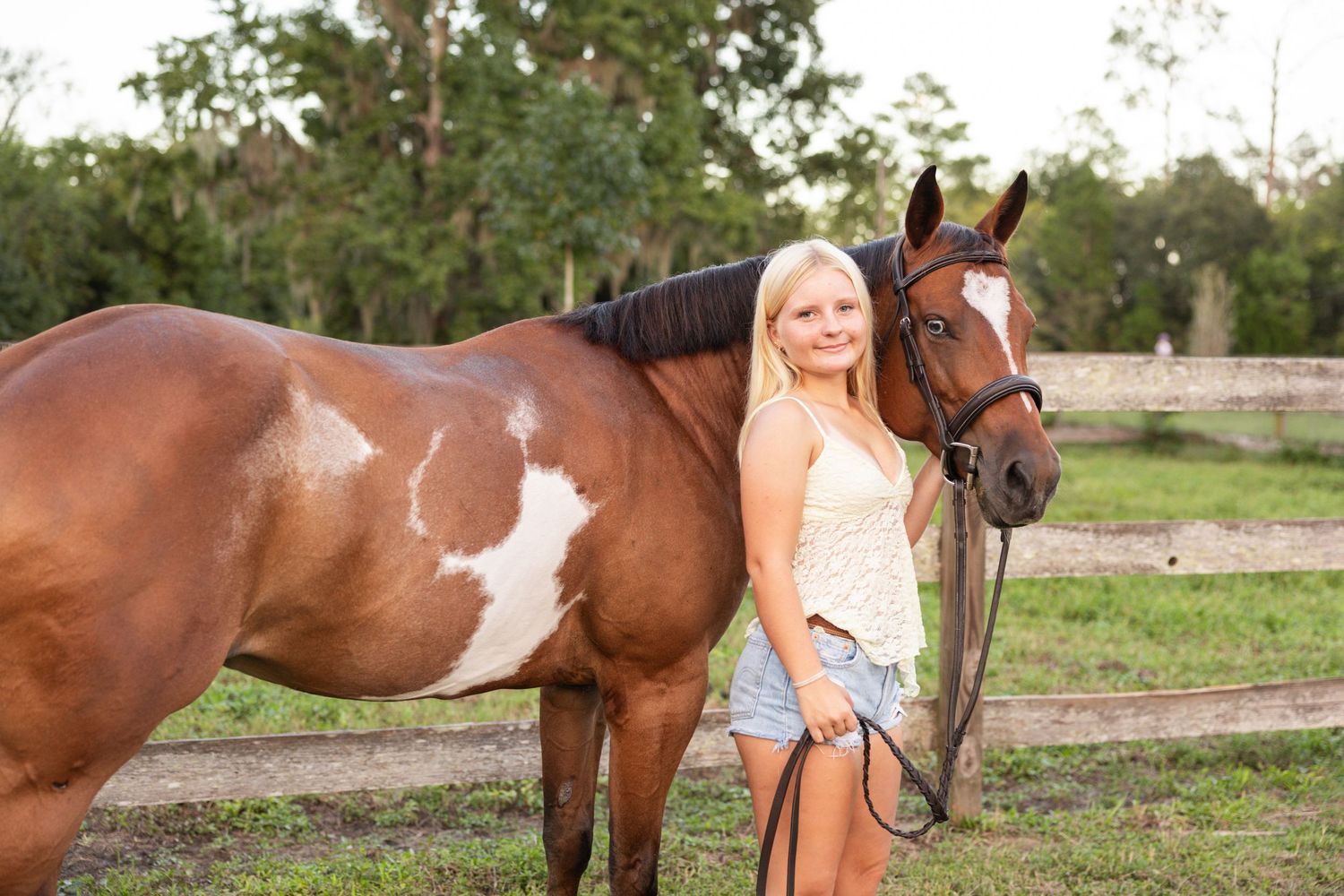 Blonde woman in denim shorts stands beside a brown and white paint horse in a green pasture.