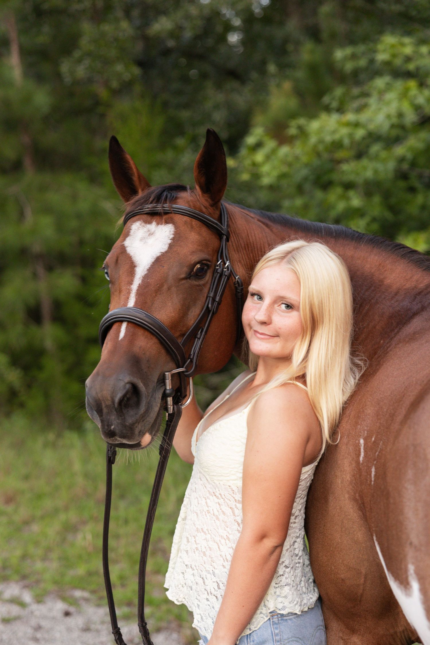 Smiling blonde girl leaning against a chestnut horse with a white blaze wearing a bridle outdoors.