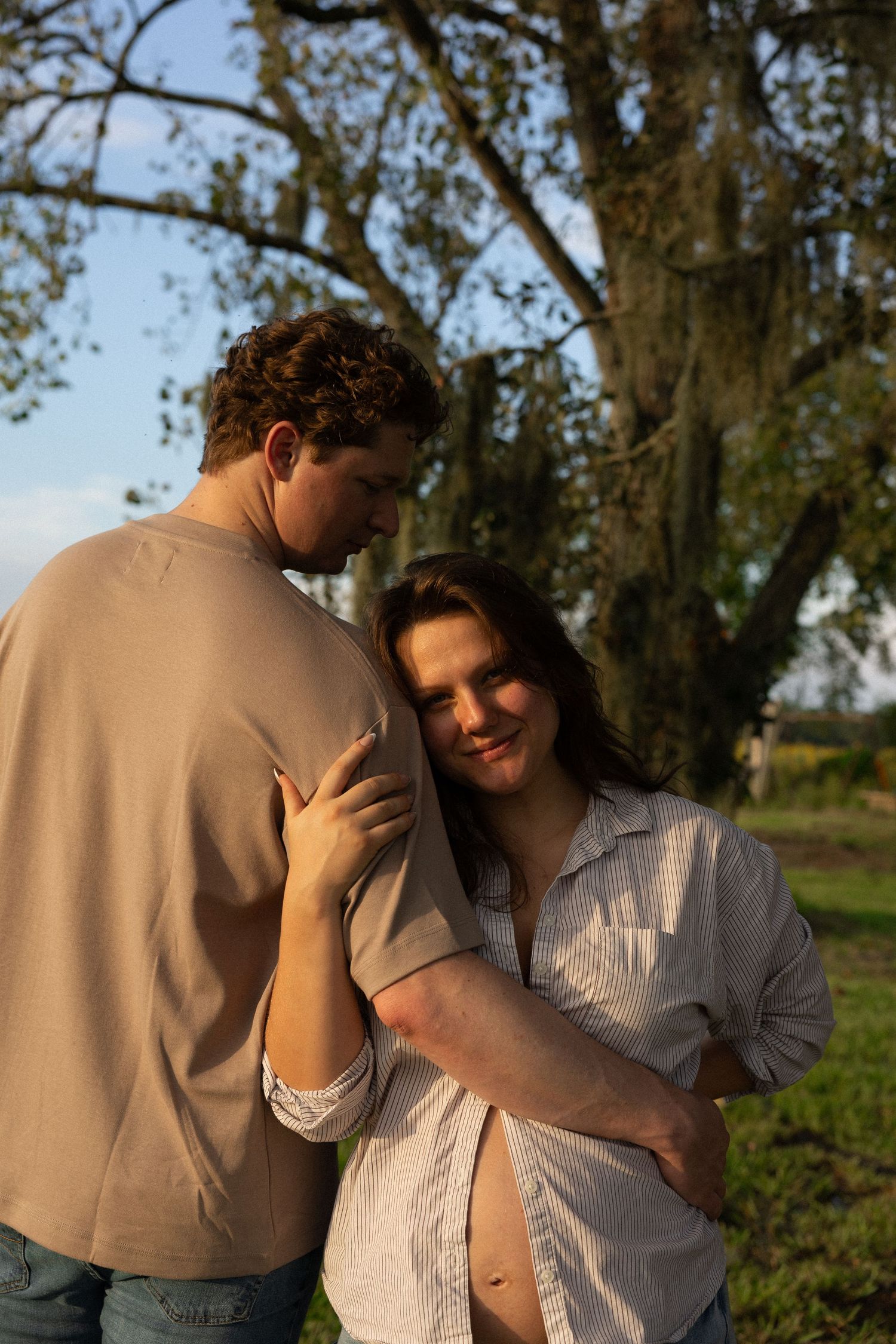 A couple embraces tenderly outdoors near a large tree in golden hour light, woman smiling softly.